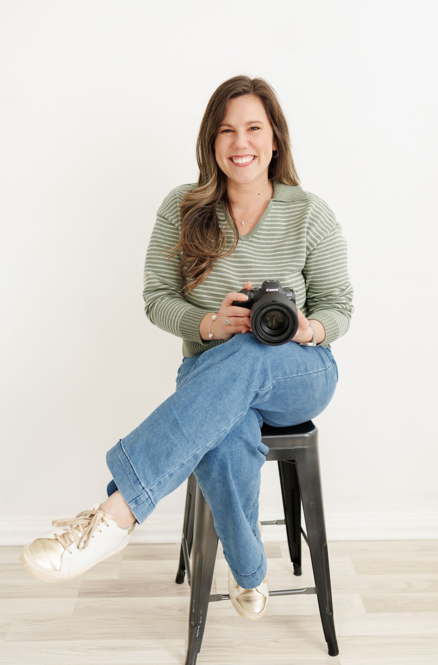 A photographer in a striped top and jeans sits casually on a black stool against a white backdrop.