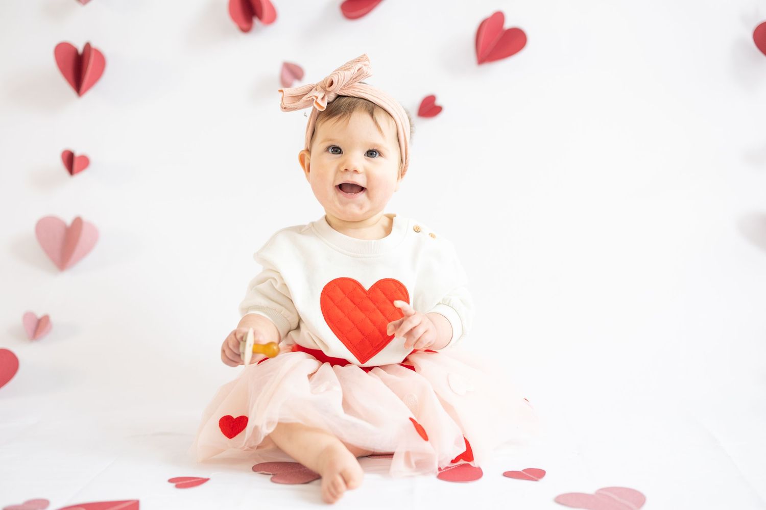 Baby in white dress holds red paper heart while sitting among floating heart decorations against white background.
