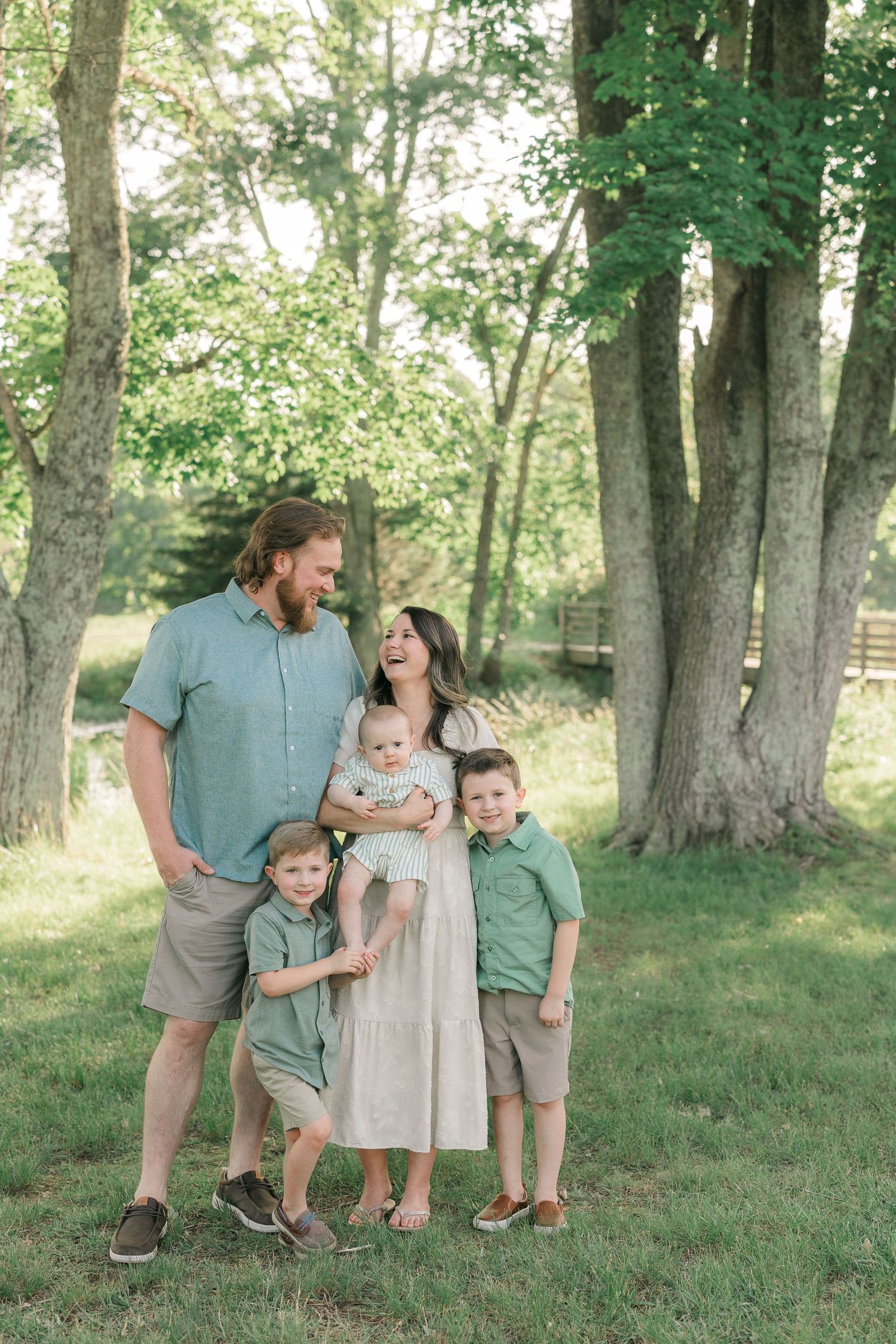 A family wearing coordinated green and beige outfits poses together in a sunlit wooded area during a photo session.