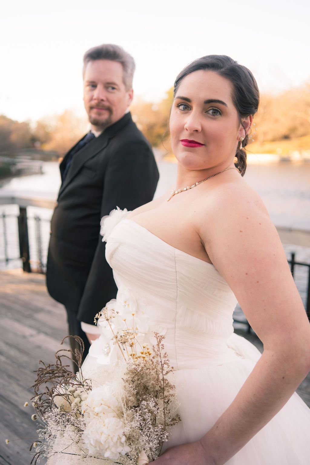 A bride in a white strapless gown poses with her groom at sunset near water.