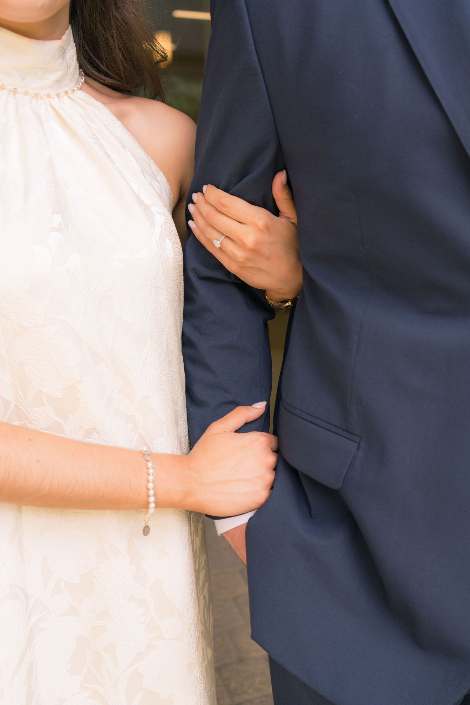 Close-up of bride in white halter gown holding groom's navy suit arm, wearing pearl bracelet and engagement ring.