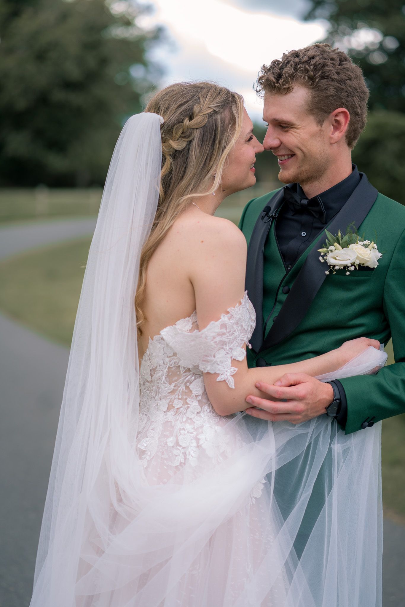A couple in wedding attire share an intimate moment outdoors with the bride wearing a lace dress and flowing veil.