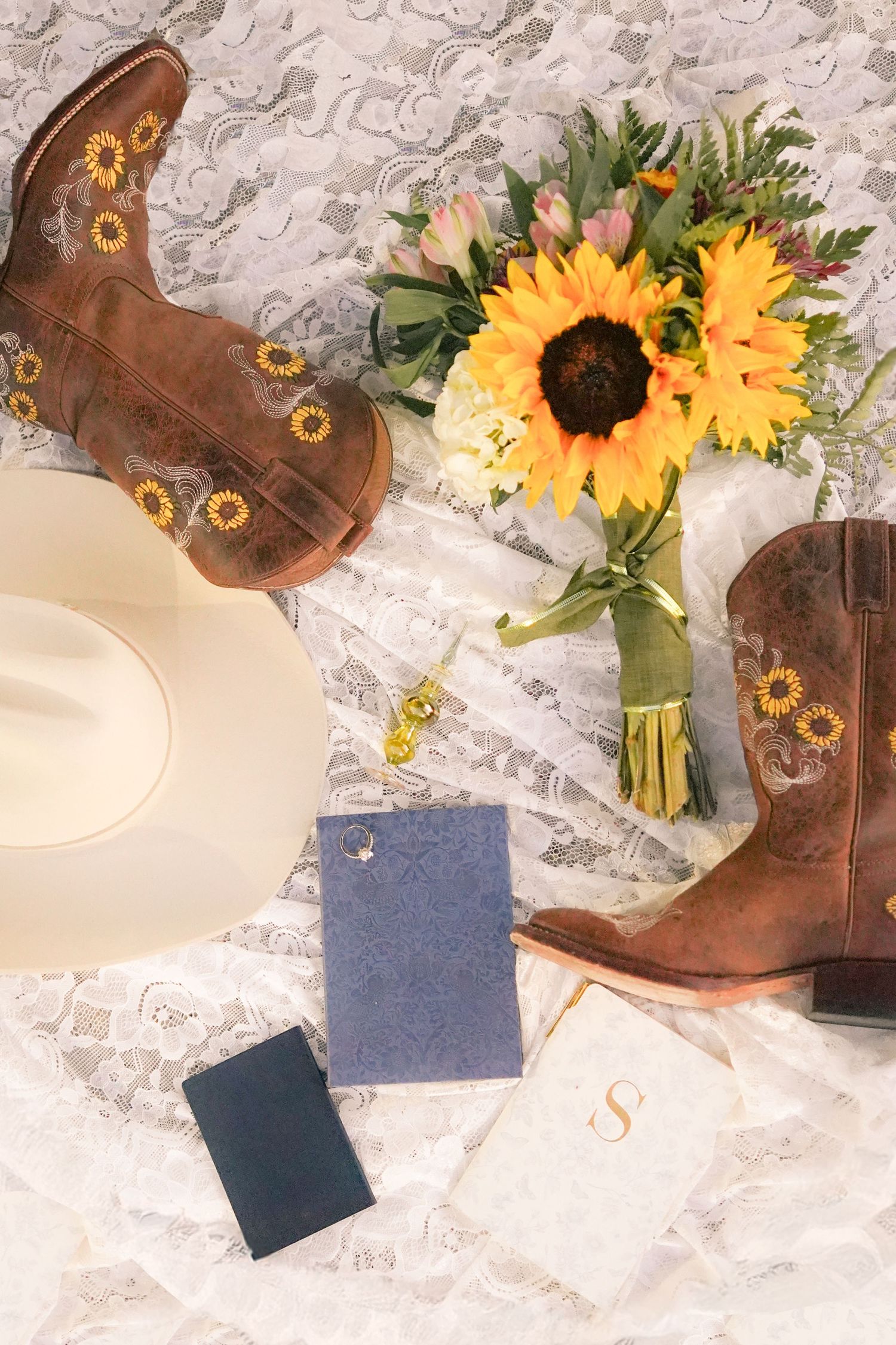 Brown cowboy boots with sunflower details displayed with a bouquet and white hat on lace.
