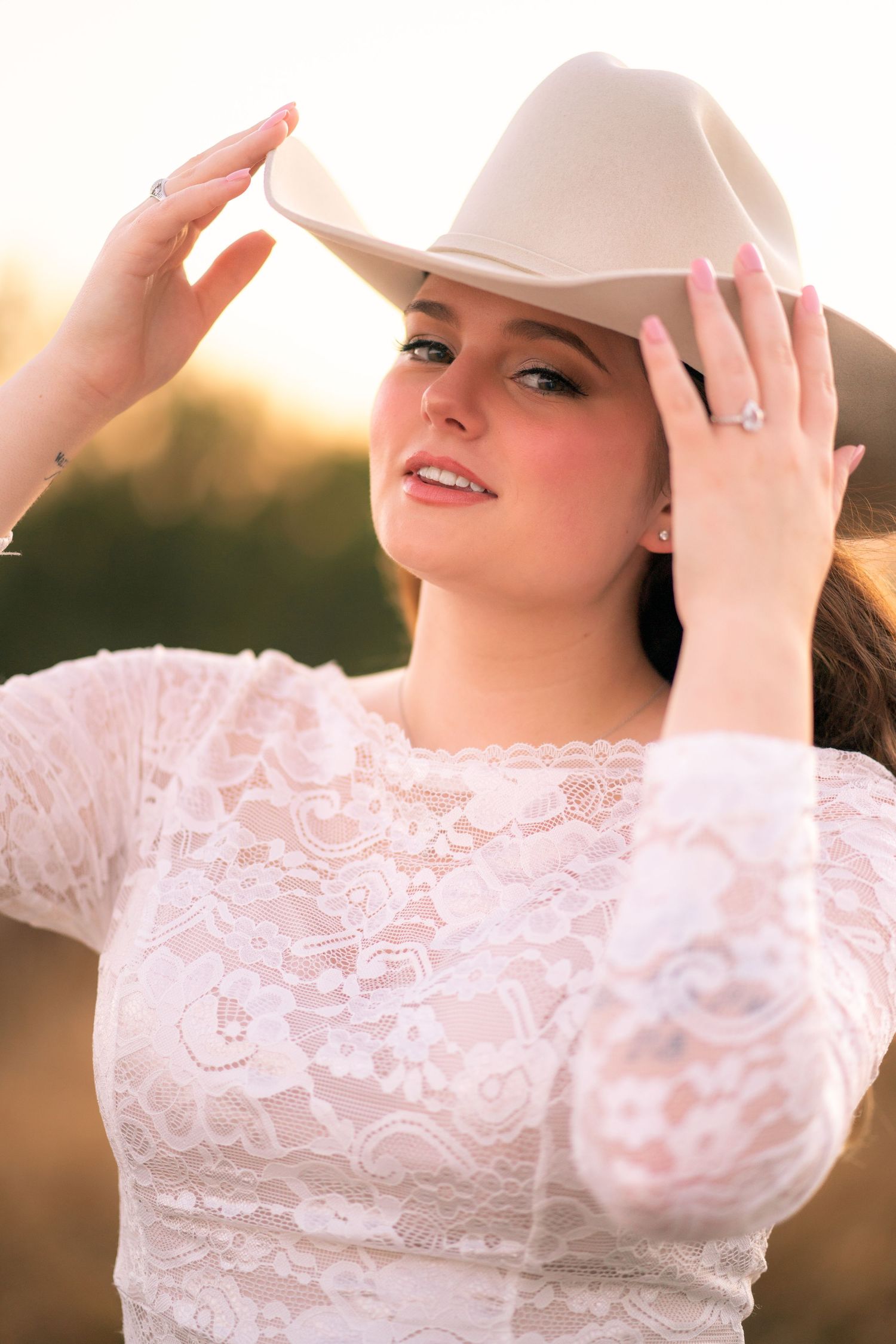 A woman in a white lace dress and beige cowboy hat poses at sunset.