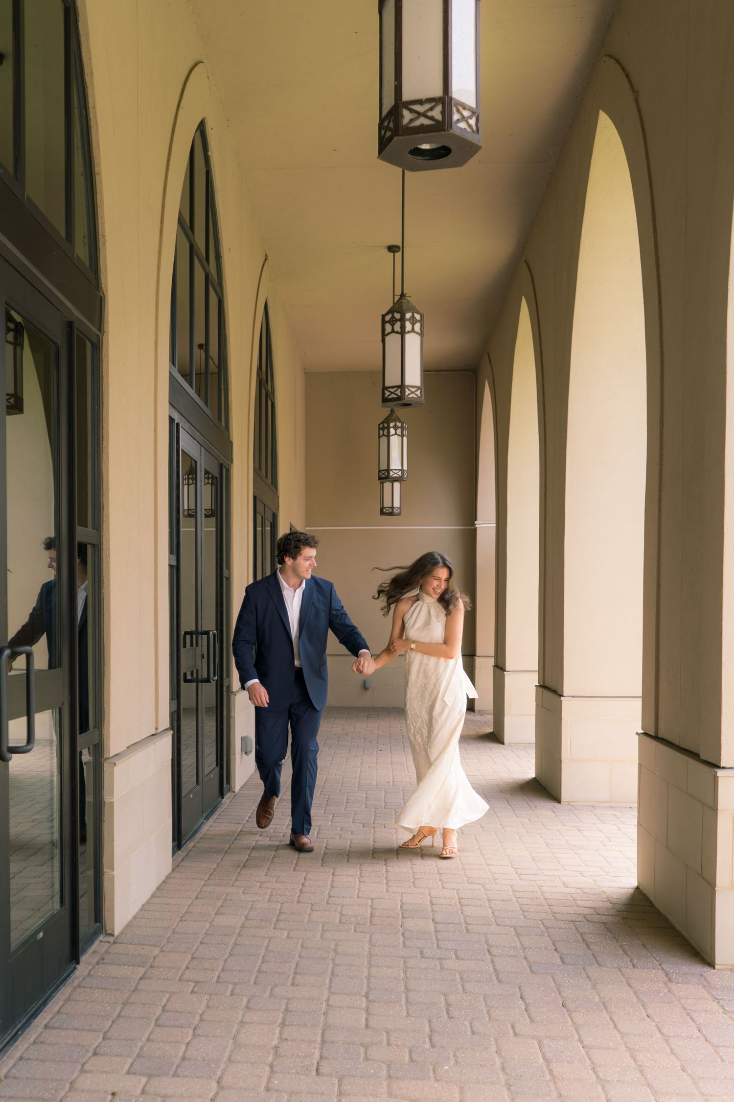 A couple in formal attire holds hands while walking through an elegant arched corridor with hanging lanterns.