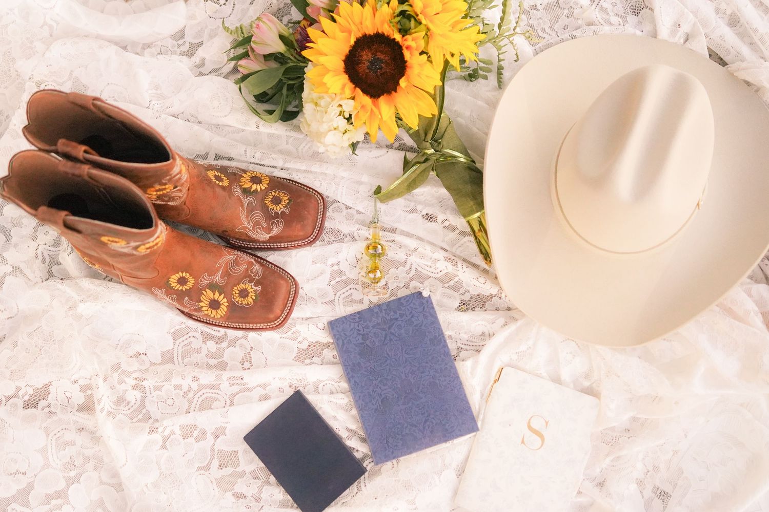 Western boots, sunflowers, and a white cowboy hat arranged on lace fabric.