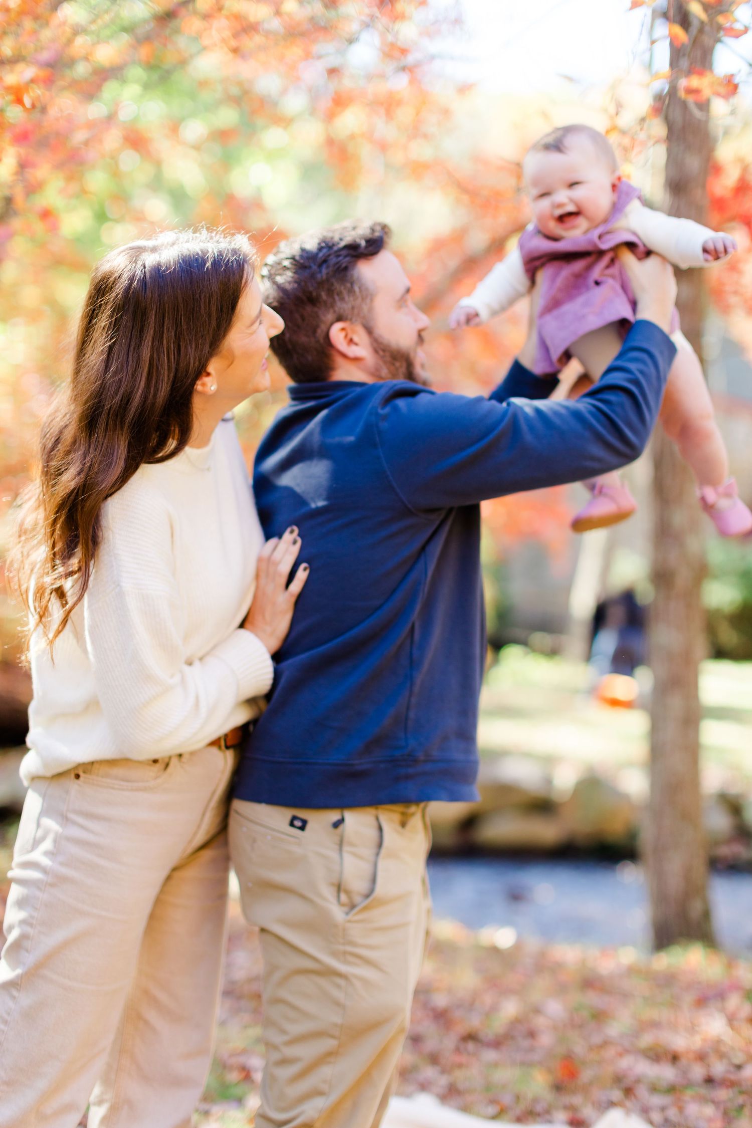 A couple and their baby playfully in a park with autumn leaves falling around them on a sunny afternoon.