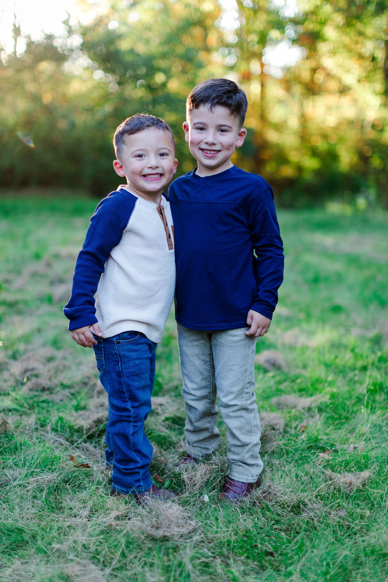 Two young children wearing casual sweaters and pants stand together smiling outdoors in a grassy area with sunlit trees behind them.