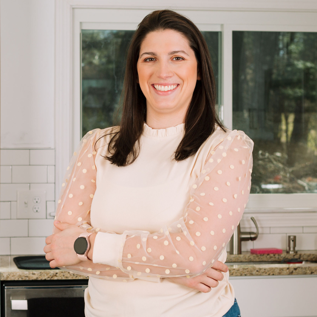 A woman in a white and peach polka dot sweater smiles while standing in a modern kitchen.