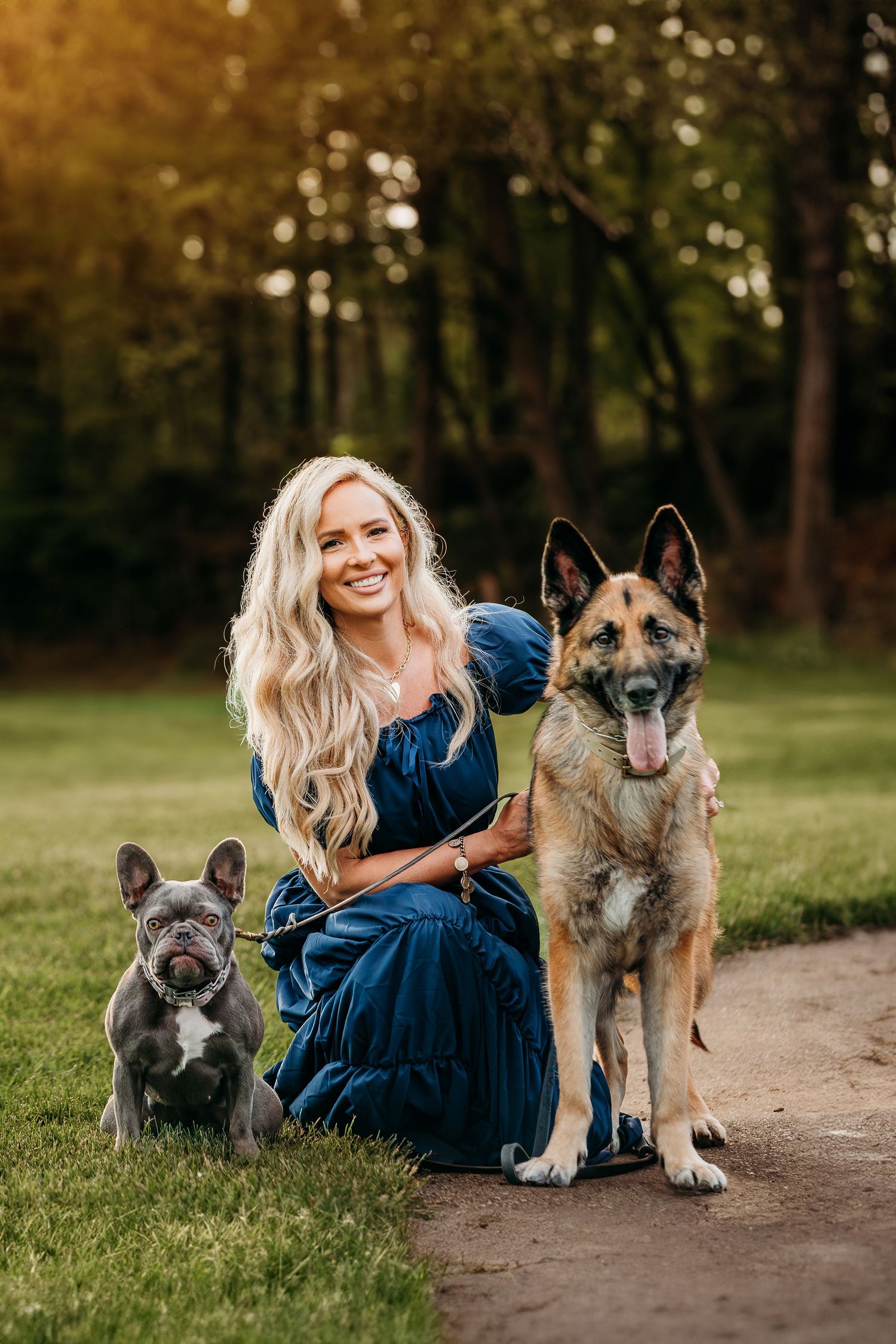 A woman in a blue dress sits on a path with two German Shepherd dogs in a park during golden hour.