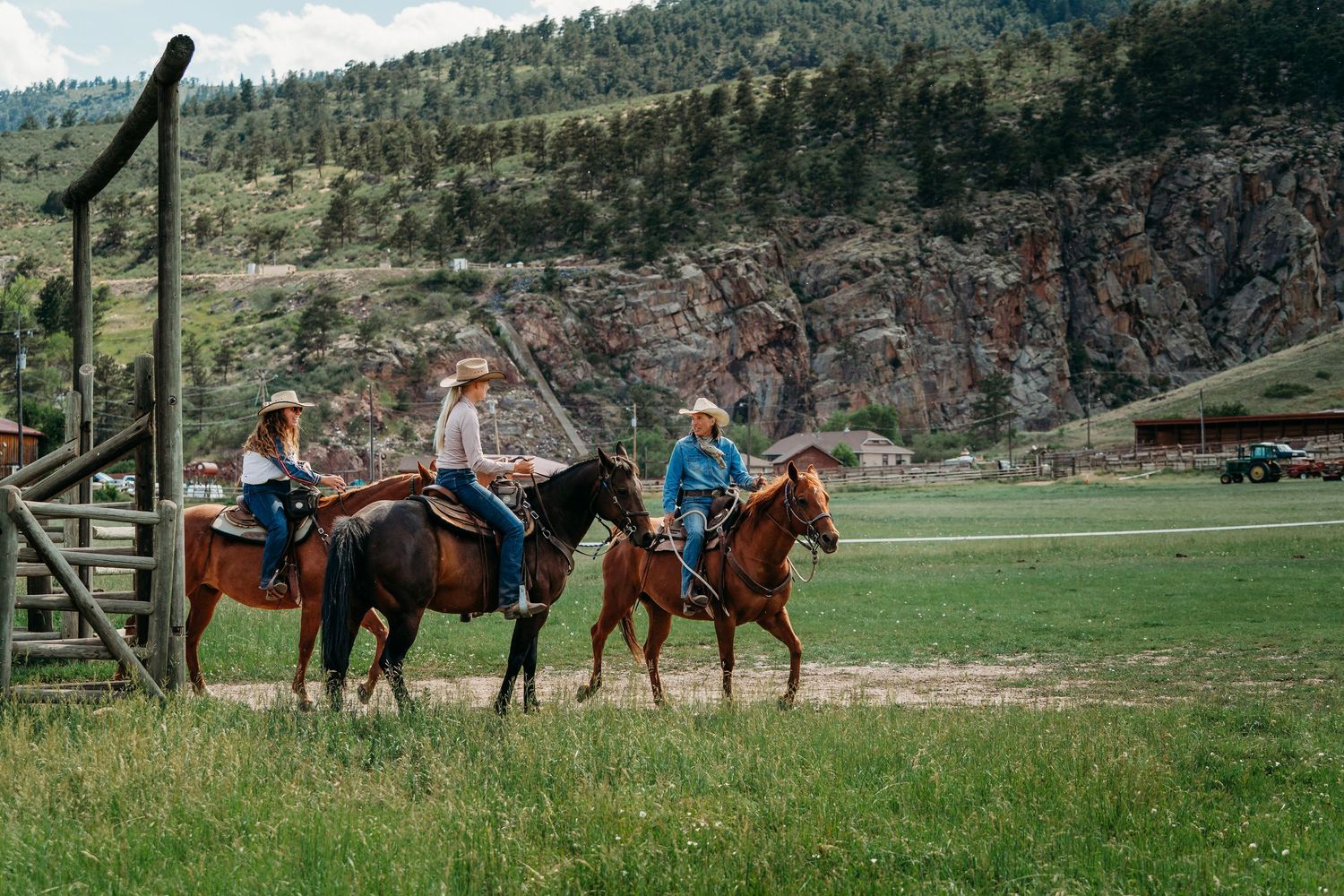 Three riders on horseback in cowboy hats traverse a green ranch valley with rocky mountain cliffs in the background.