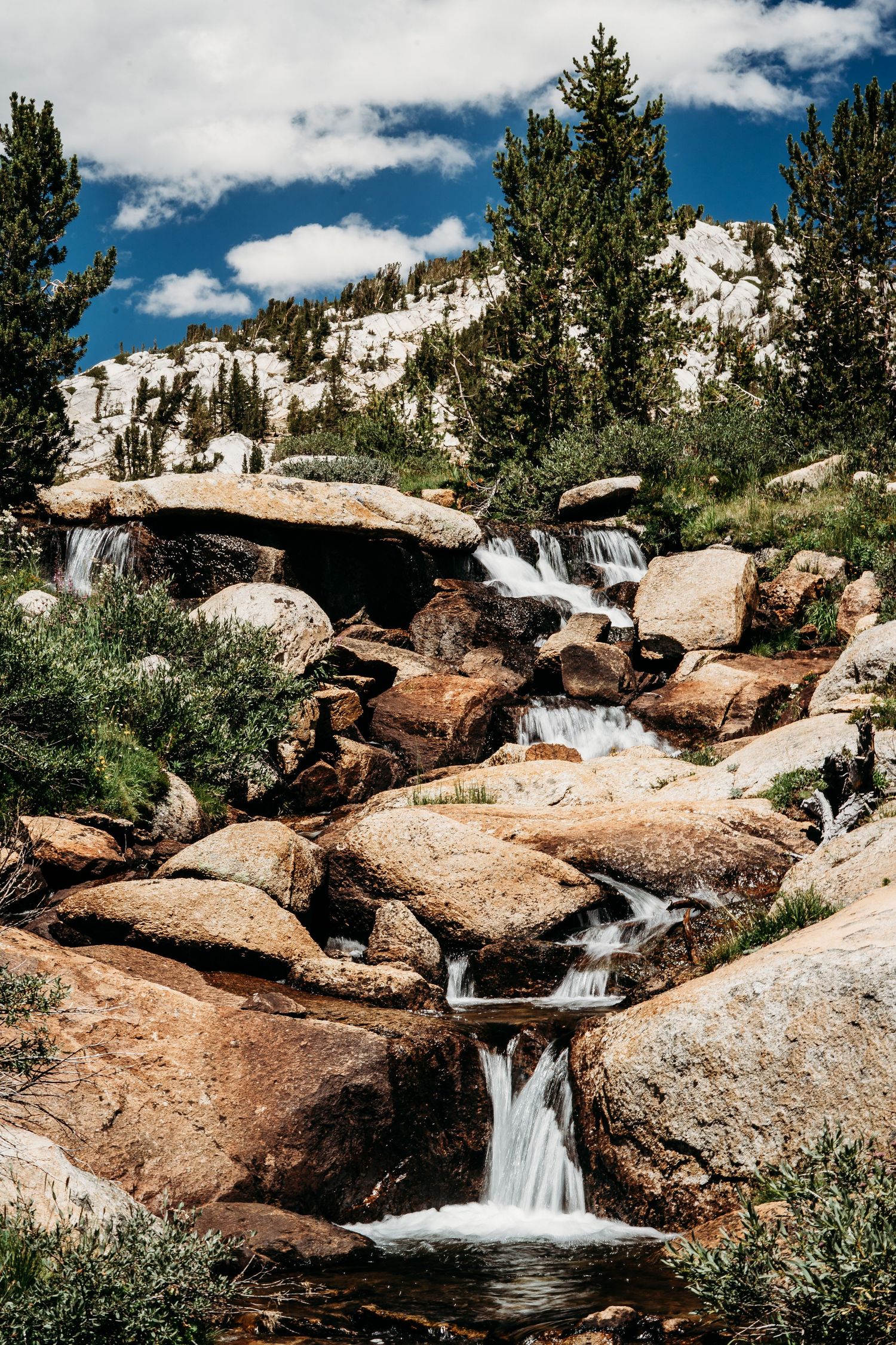 Cascading waterfall flows over rocky terrain with snow-capped mountains in the background.