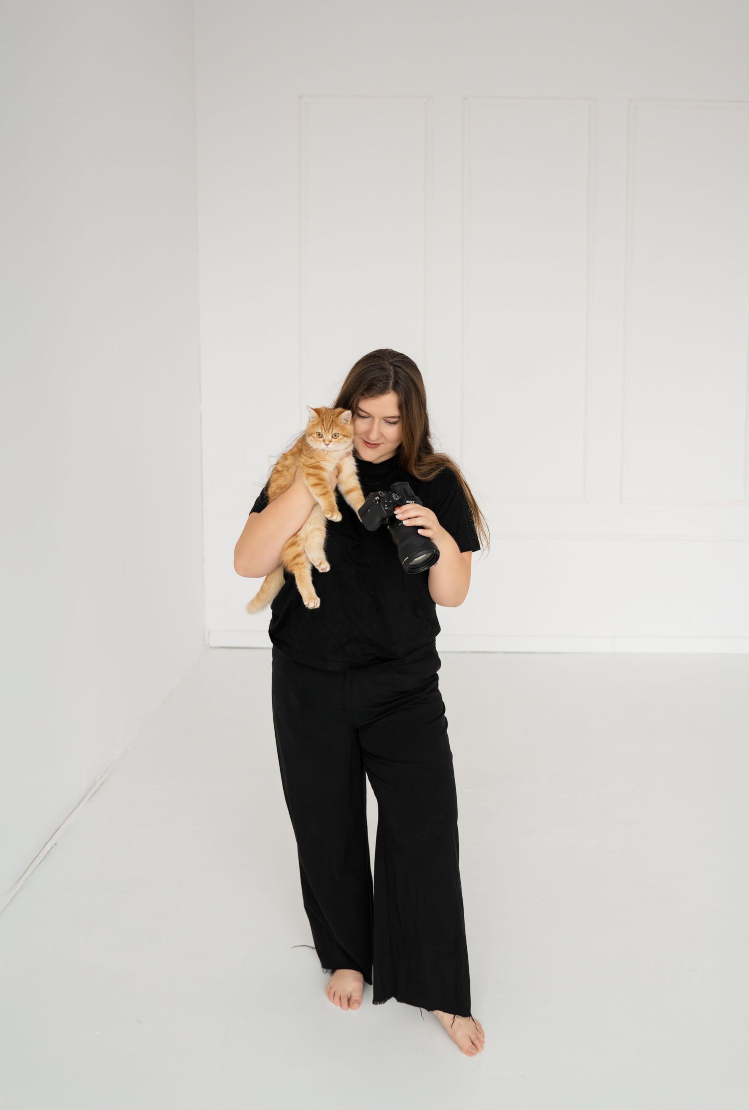Person in black outfit holding ginger cat in minimalist white studio setting.