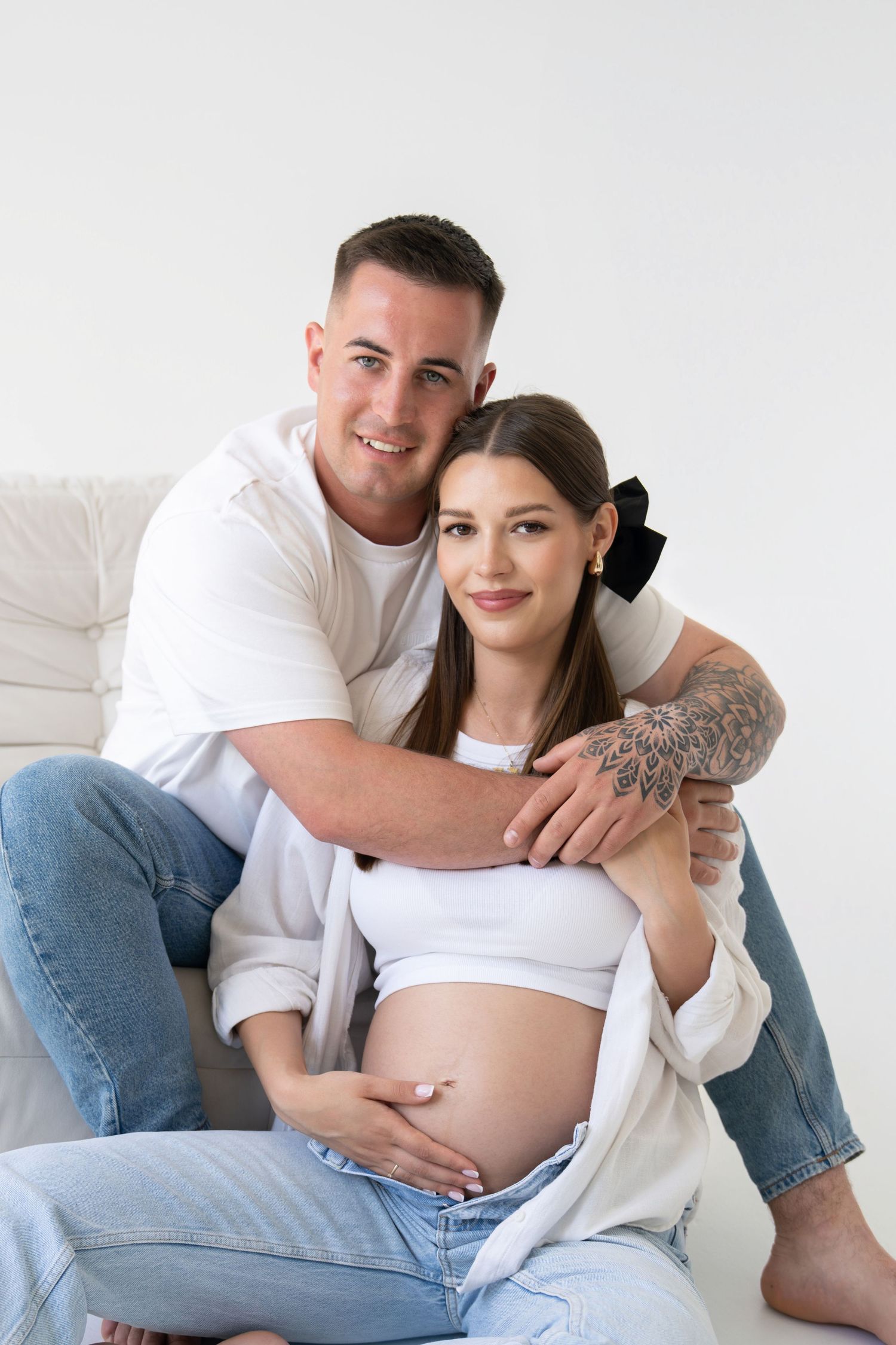 A couple in white shirts and jeans embraces during a maternity photo session against a light background.