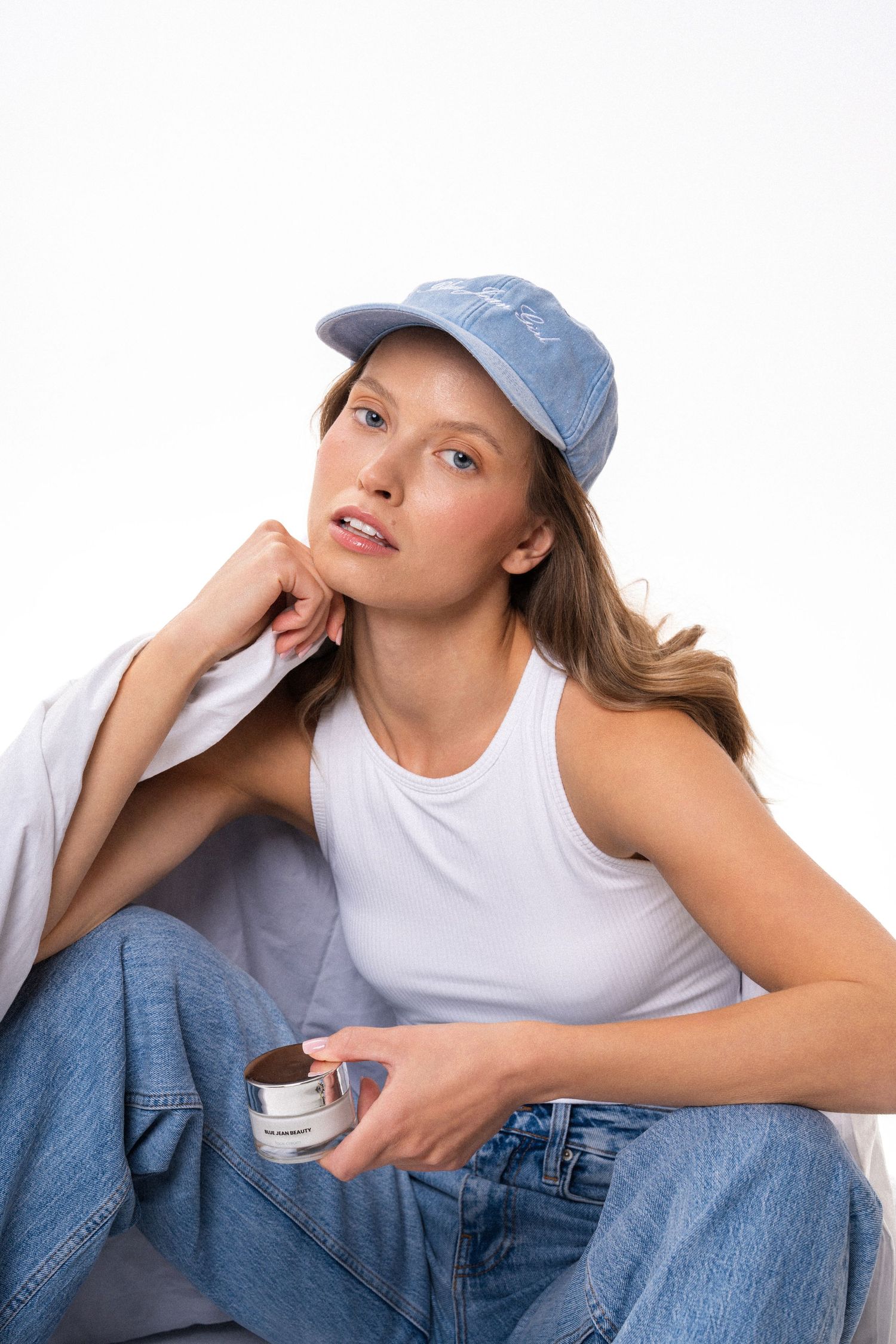 Young woman in blue cap, white tank top, and jeans sitting casually holding a skincare product jar.