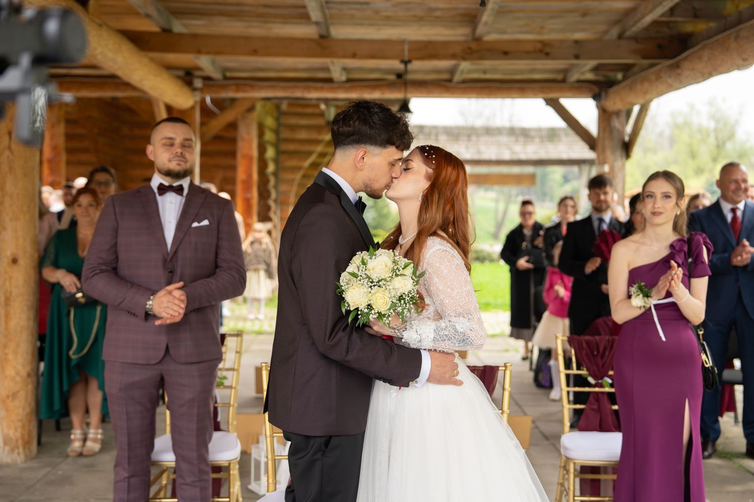 Bride and groom share first kiss at outdoor wedding ceremony, guests applauding in background.