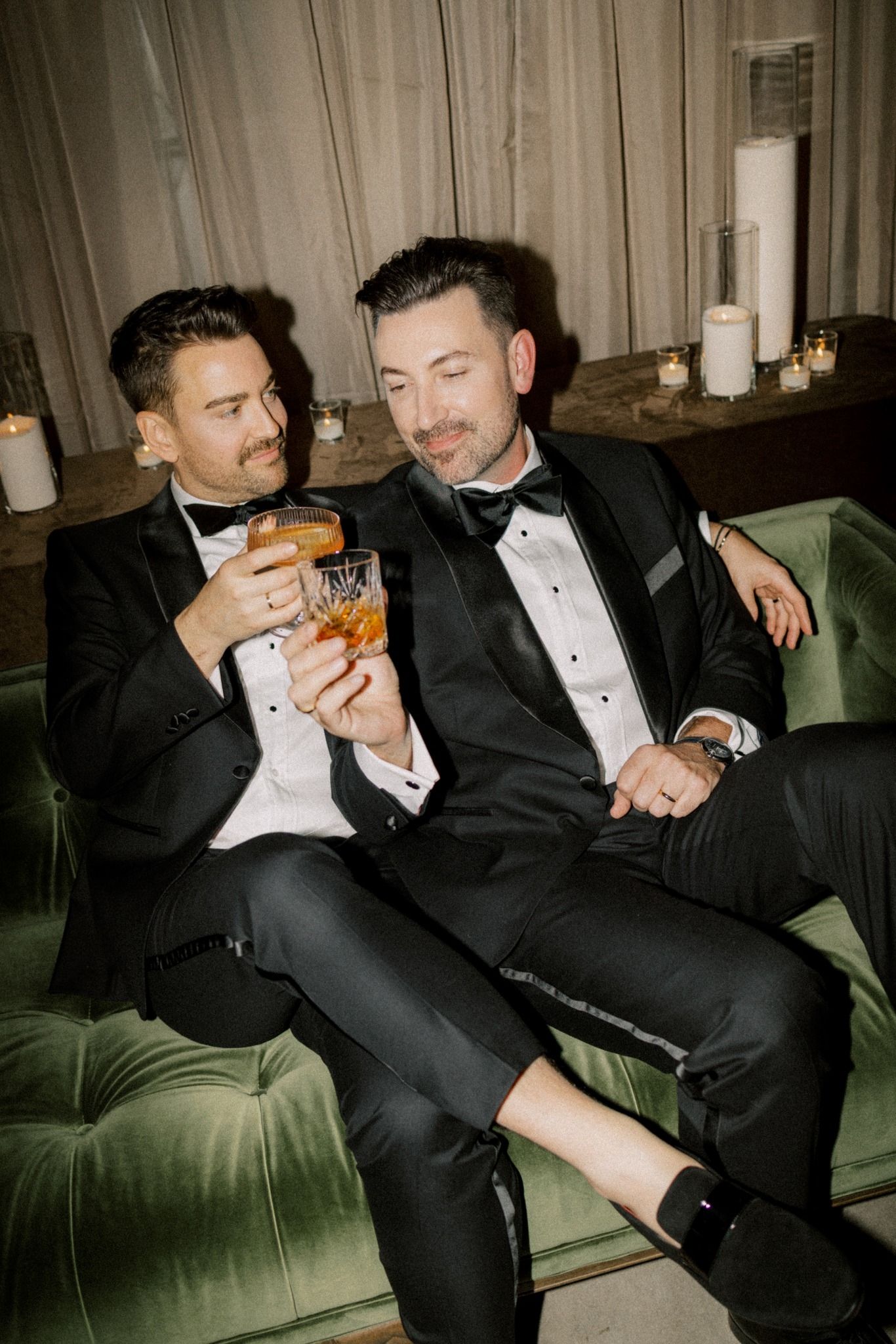 Two men in tuxedos toasting with whiskey glasses on a green velvet sofa at a formal event.