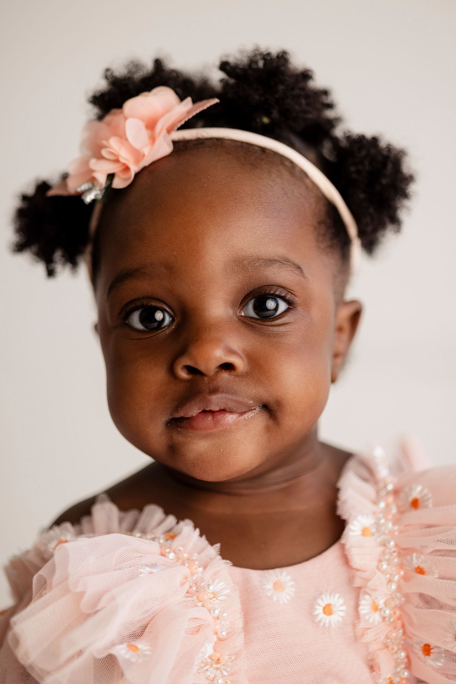 A young child wearing a pink dress and floral headband with hair styled in puffs.