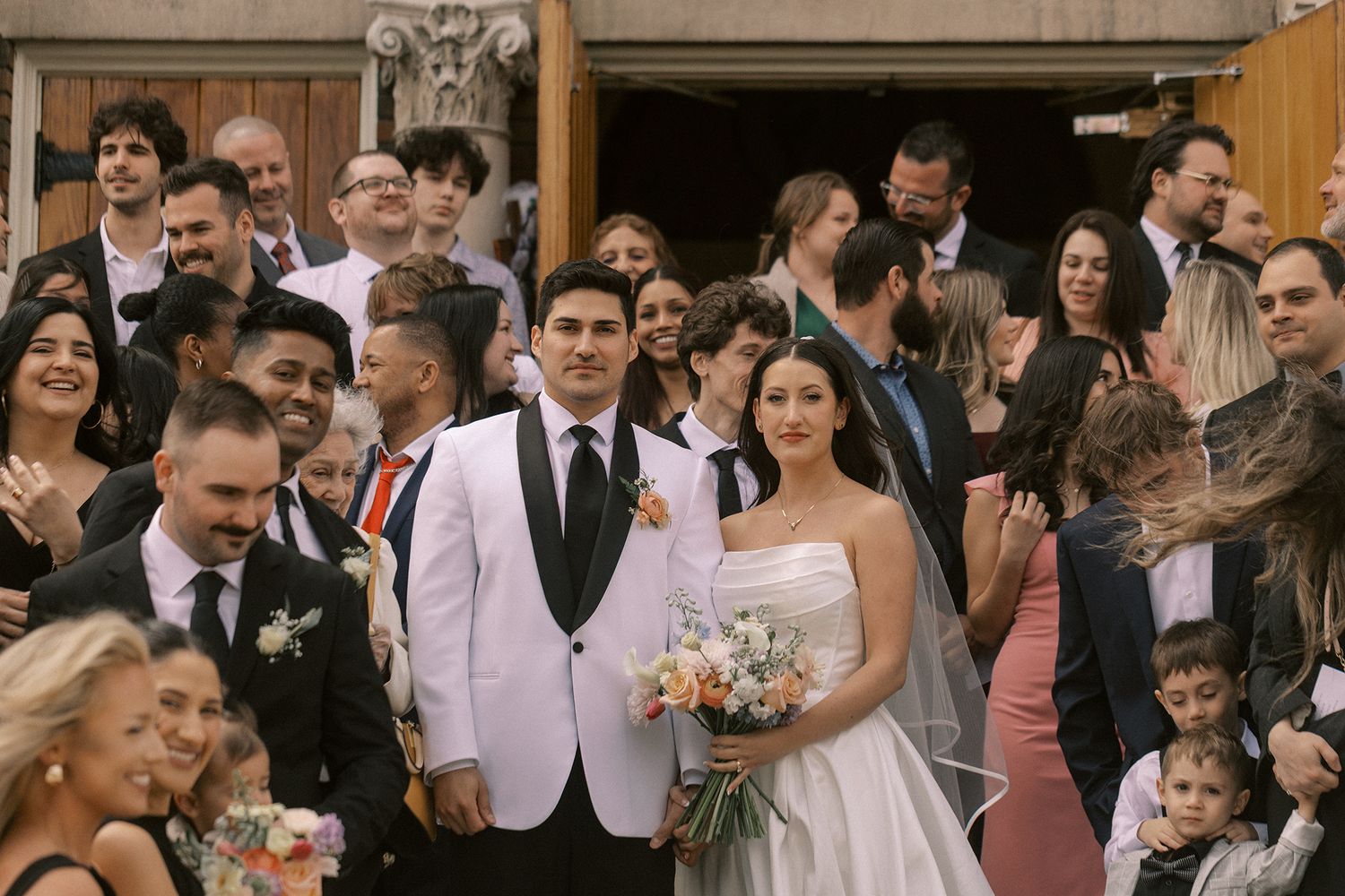 Newlyweds pose with wedding party and guests outside church entrance after ceremony.