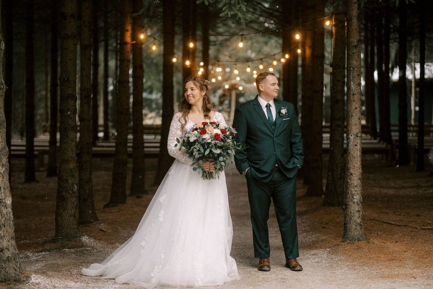 A bride and groom pose together in a magical forest setting lit by string lights.