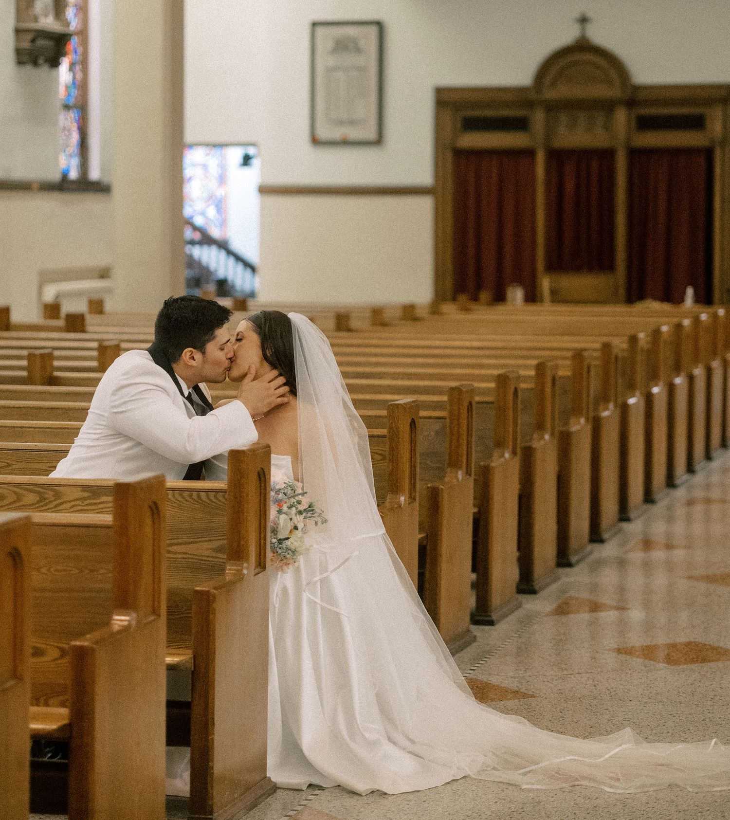 A bride and groom share their first kiss in a traditional church setting with wooden pews.