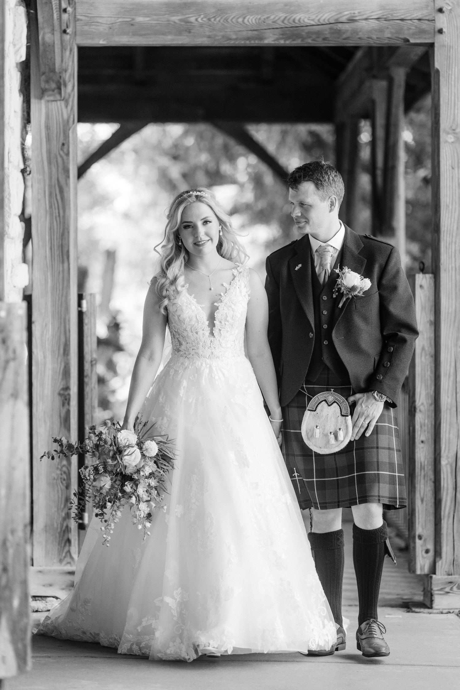 A bride in a white lace gown and groom in traditional Scottish kilt pose together under a covered walkway.