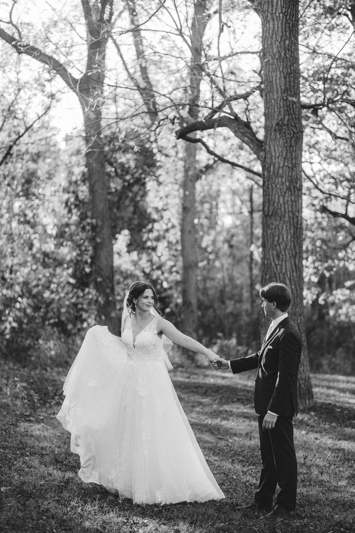 Black and white photo of couple dancing together on grassy lawn surrounded by trees.
