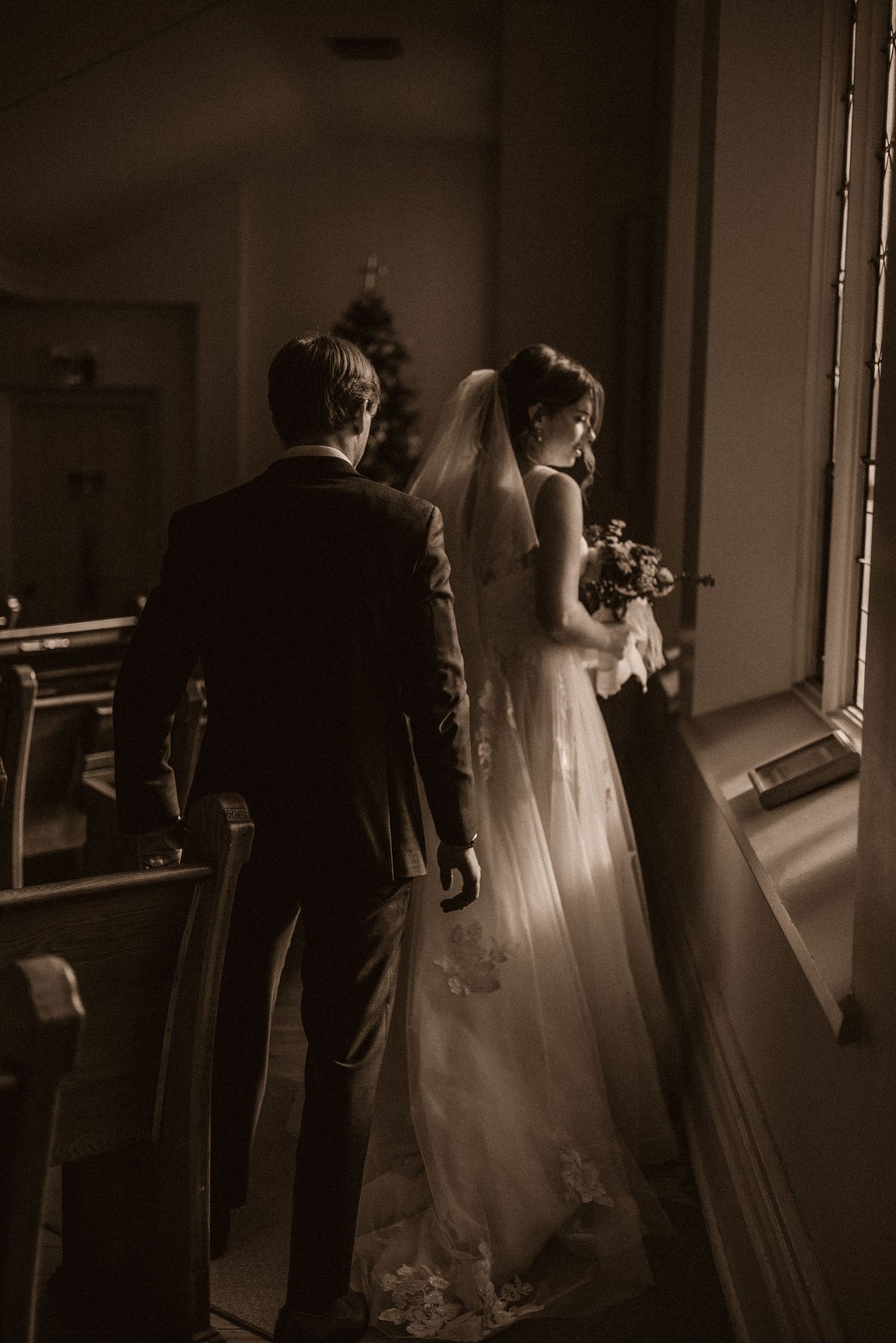 Dramatic sepia photo of couple by window in formal wedding attire.