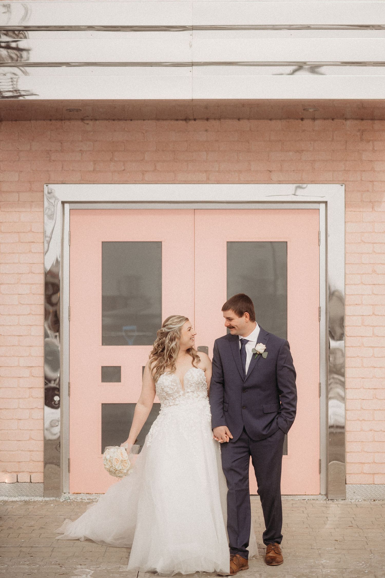 A bride and groom pose in front of a pink door for their wedding portrait.