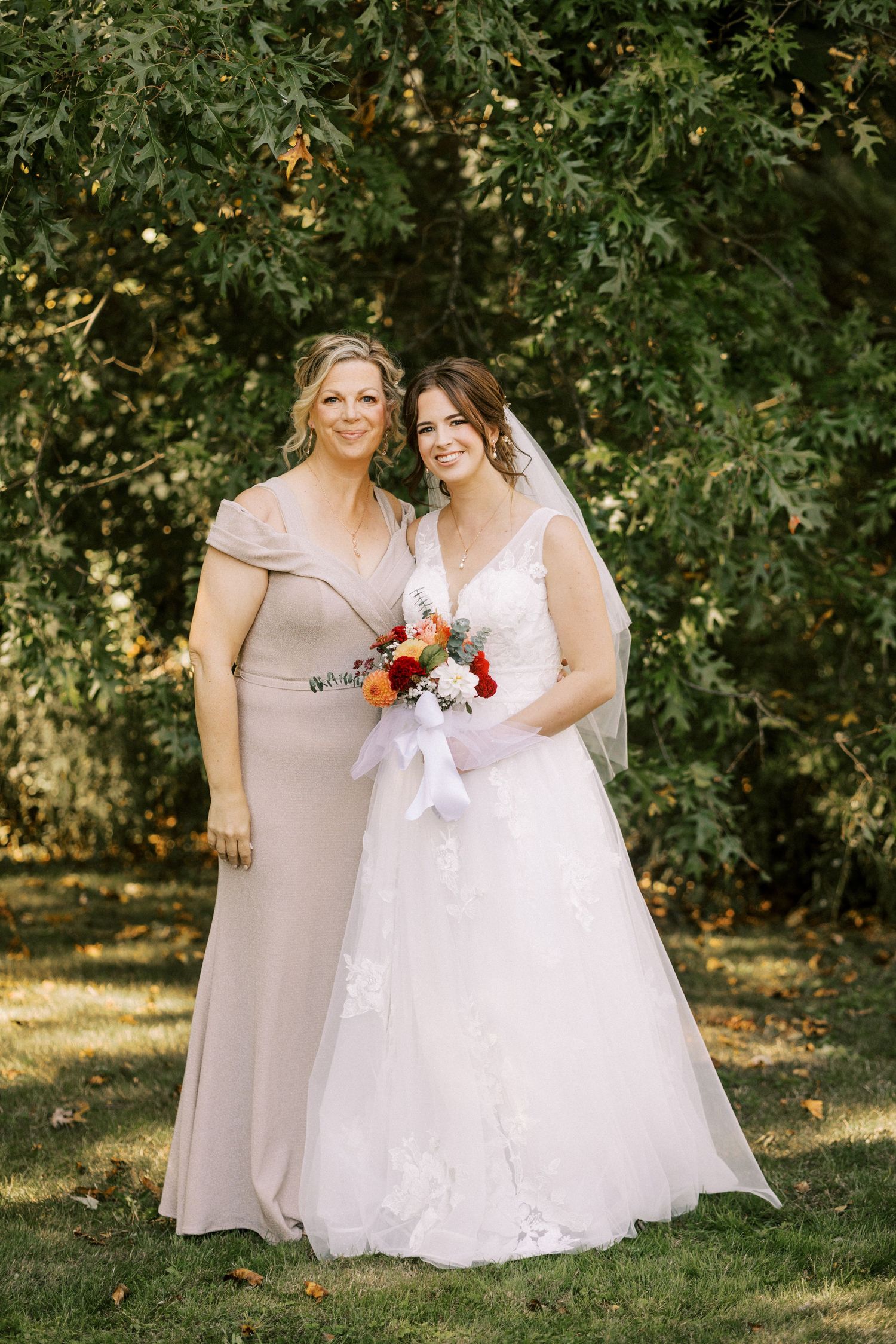 Two people in formal attire pose together outdoors holding autumn-colored bouquet.