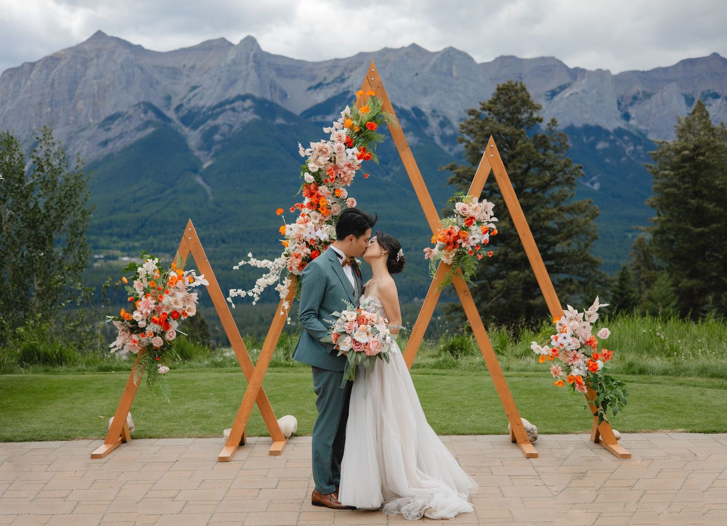 Couple kissing at mountain wedding altar with geometric wooden arch and floral arrangements in Canmore, Alberta.