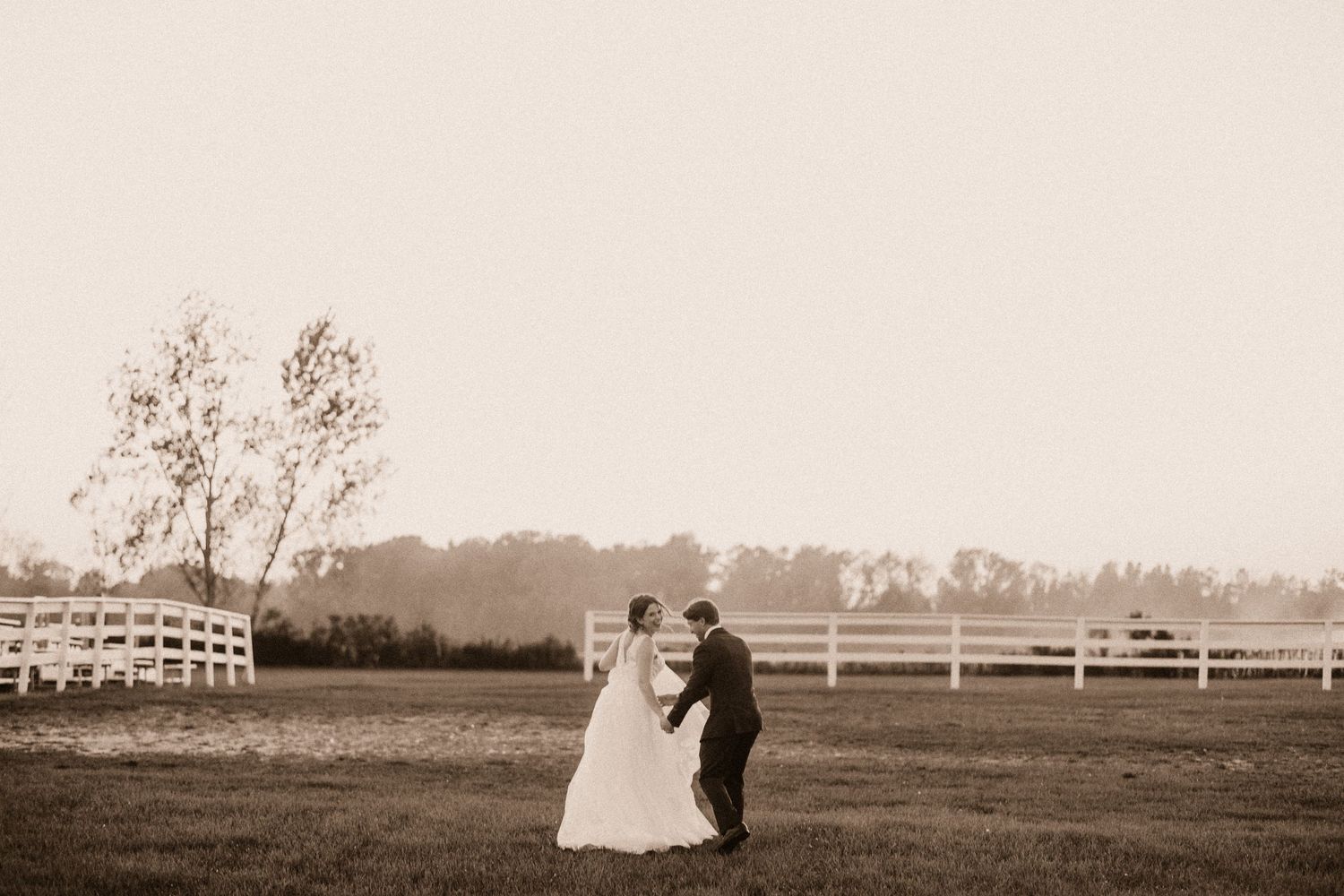 A sepia-toned wedding photograph showing a couple embracing in a pastoral field with white fences.