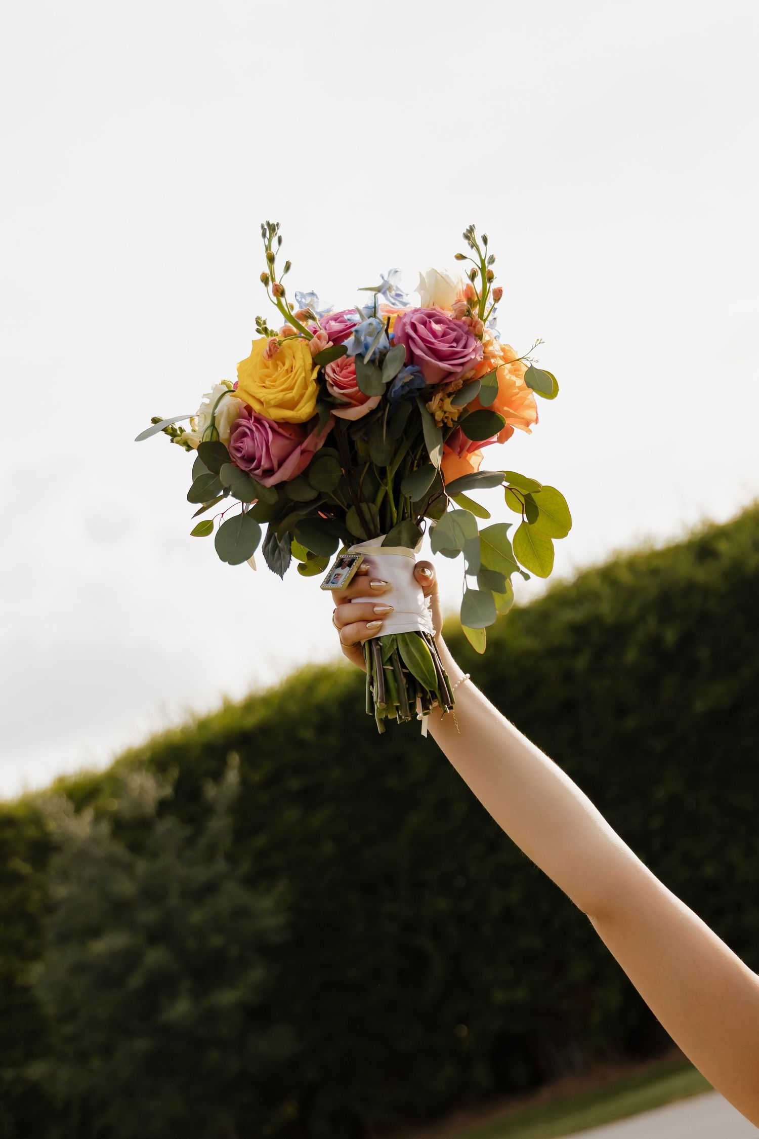 Hand raised high holding a vibrant wedding bouquet with pink, yellow, orange, and purple roses against a cloudy sky.