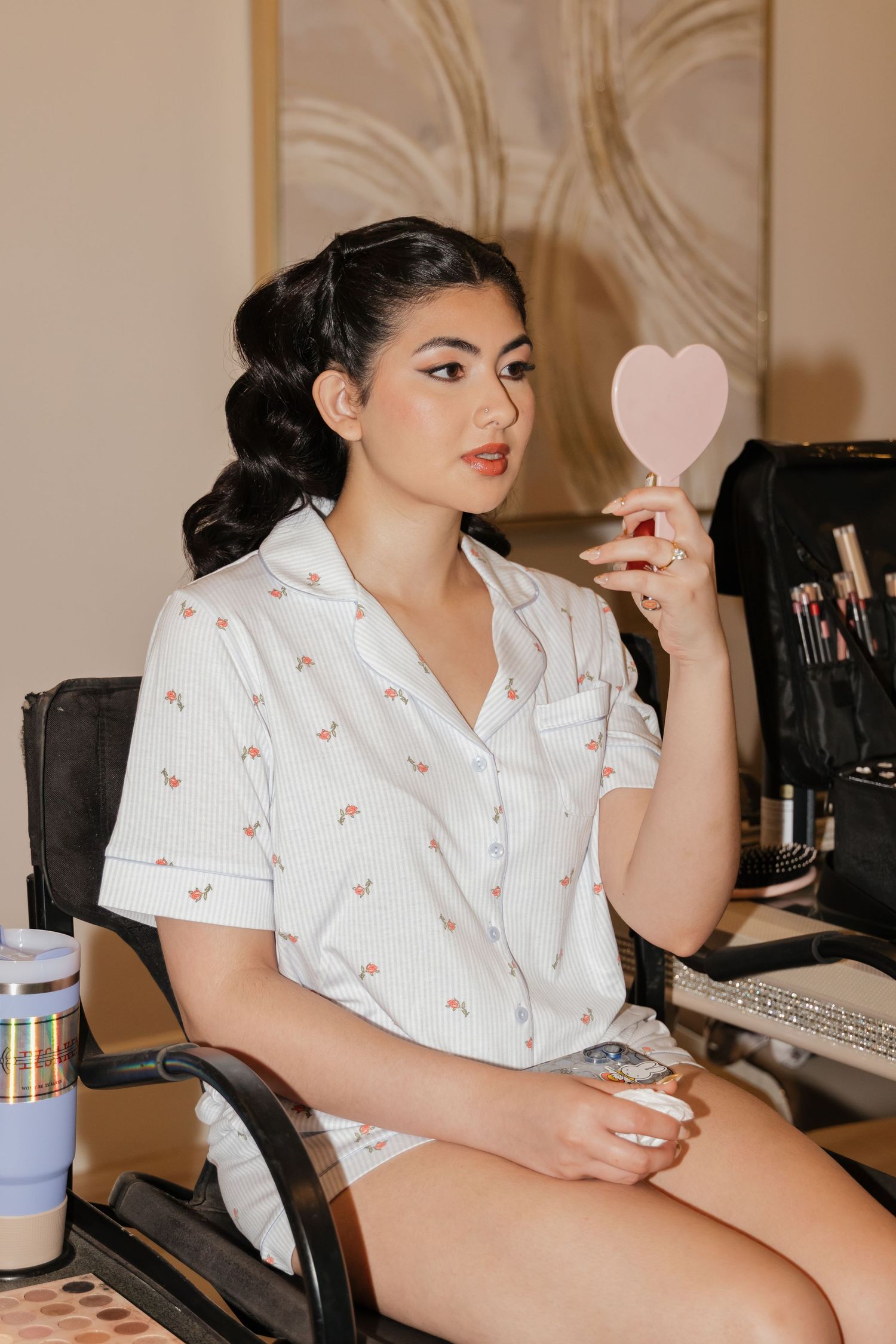 Young woman in floral pajamas holds a pink heart-shaped mirror at a vanity table with makeup brushes.