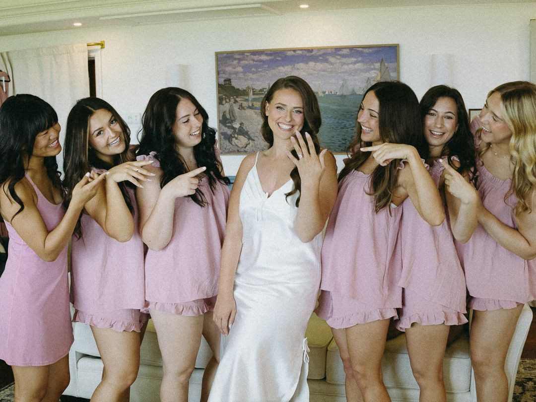 Bride in white gown surrounded by bridesmaids in pink dresses, smiling and showing off engagement ring indoors.
