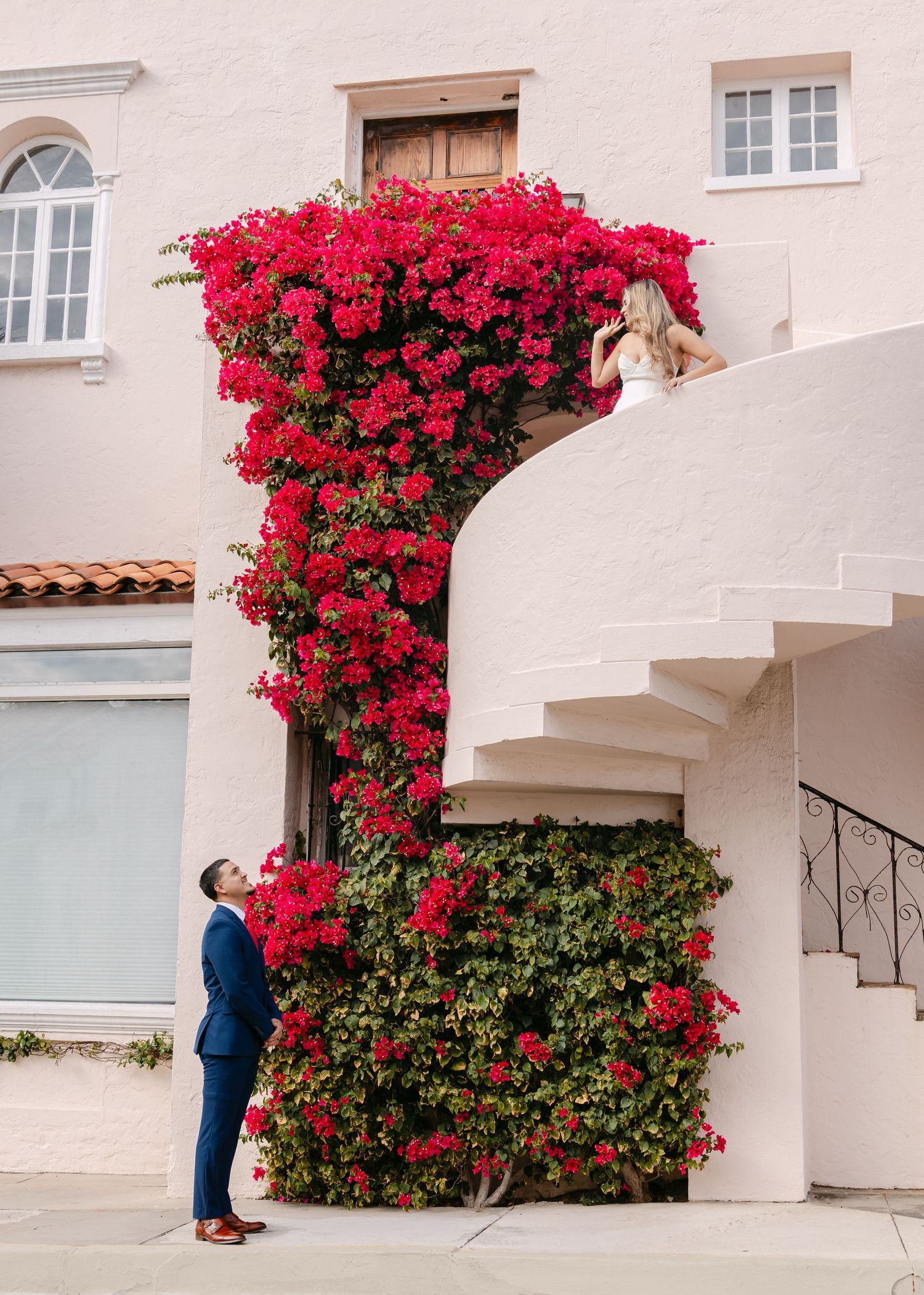 Couple poses dramatically on white spiral staircase adorned with vibrant red bougainvillea blooms.
