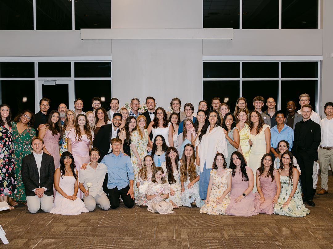 Large group wedding photo with bride, groom, and guests gathered indoors at the reception venue.