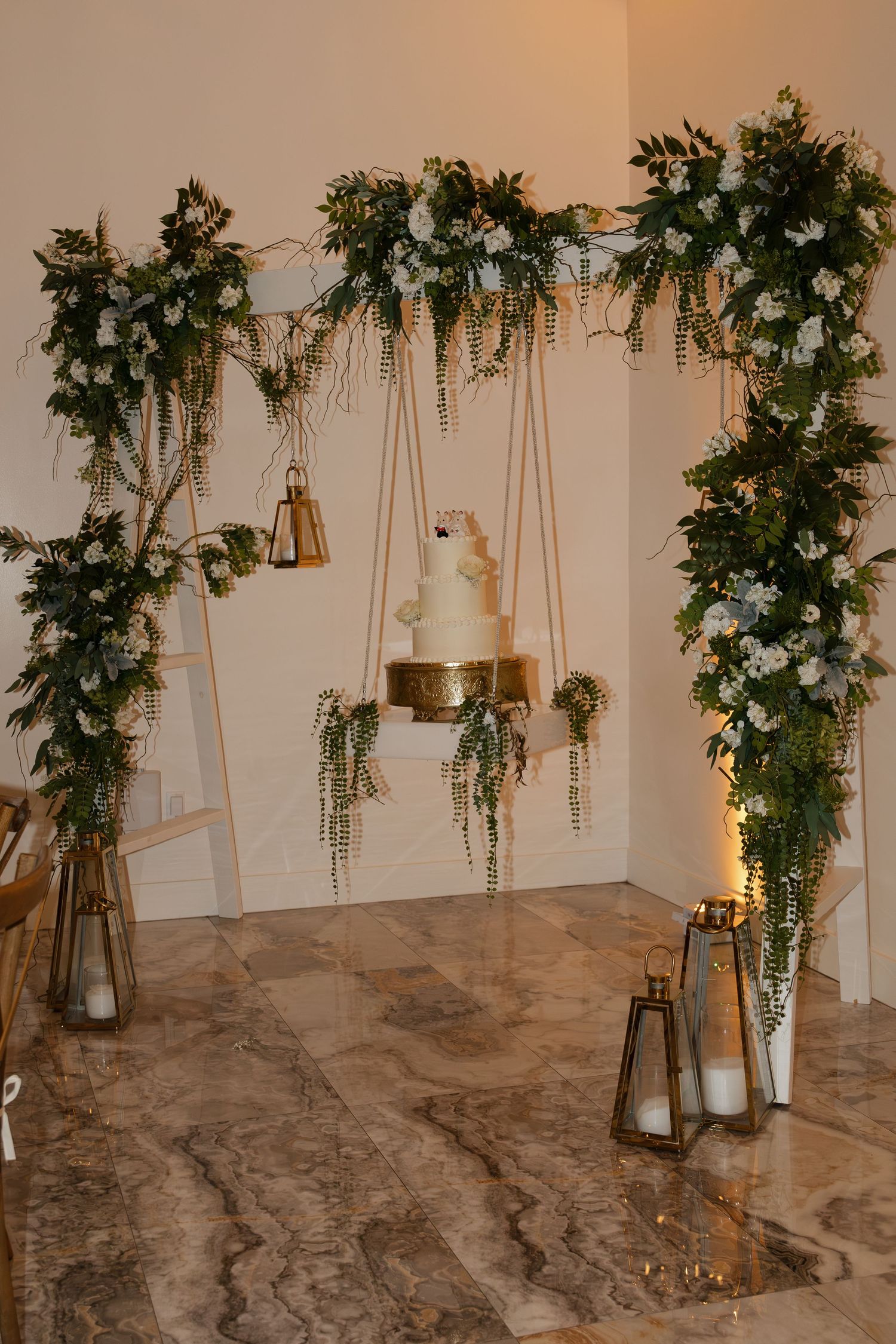 Elegant wedding cake display suspended from a floral arch with greenery and white flowers, gold lanterns on marble floor.