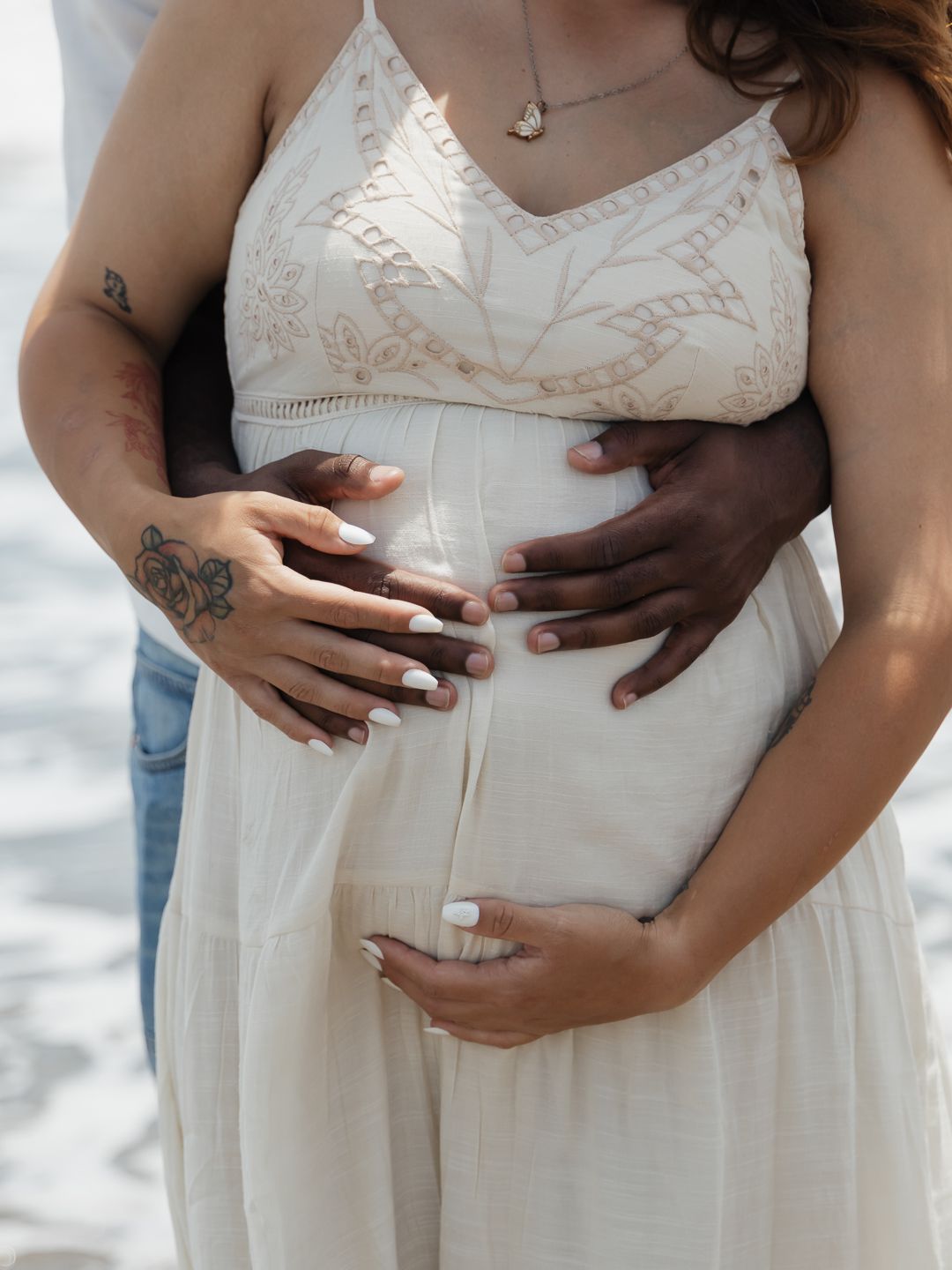 Pregnant woman in white dress with partner's hands cradling baby bump near ocean beach maternity photo.