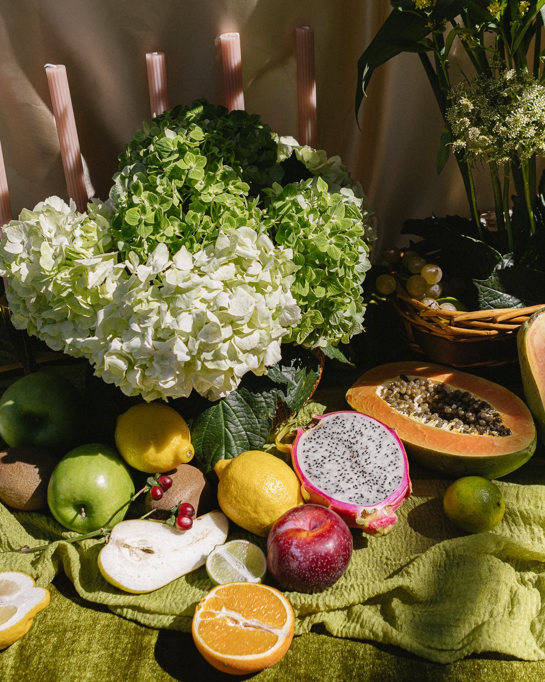 A vibrant still life display of tropical fruits, green apples, lemons, and white hydrangeas on a green cloth.