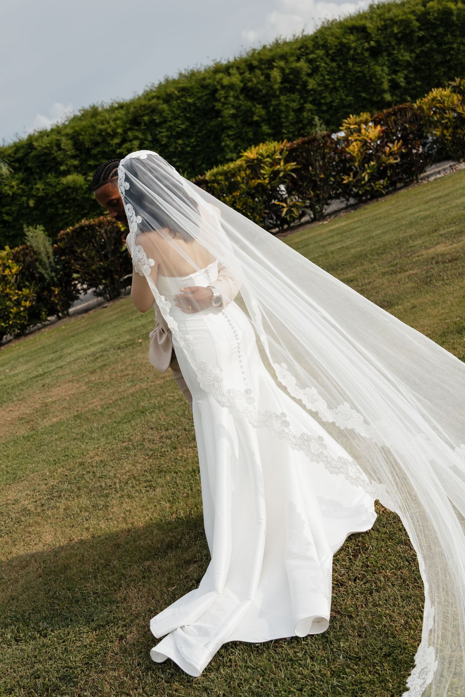 Bride in a white mermaid gown with long flowing lace veil blowing in the wind on a green lawn.