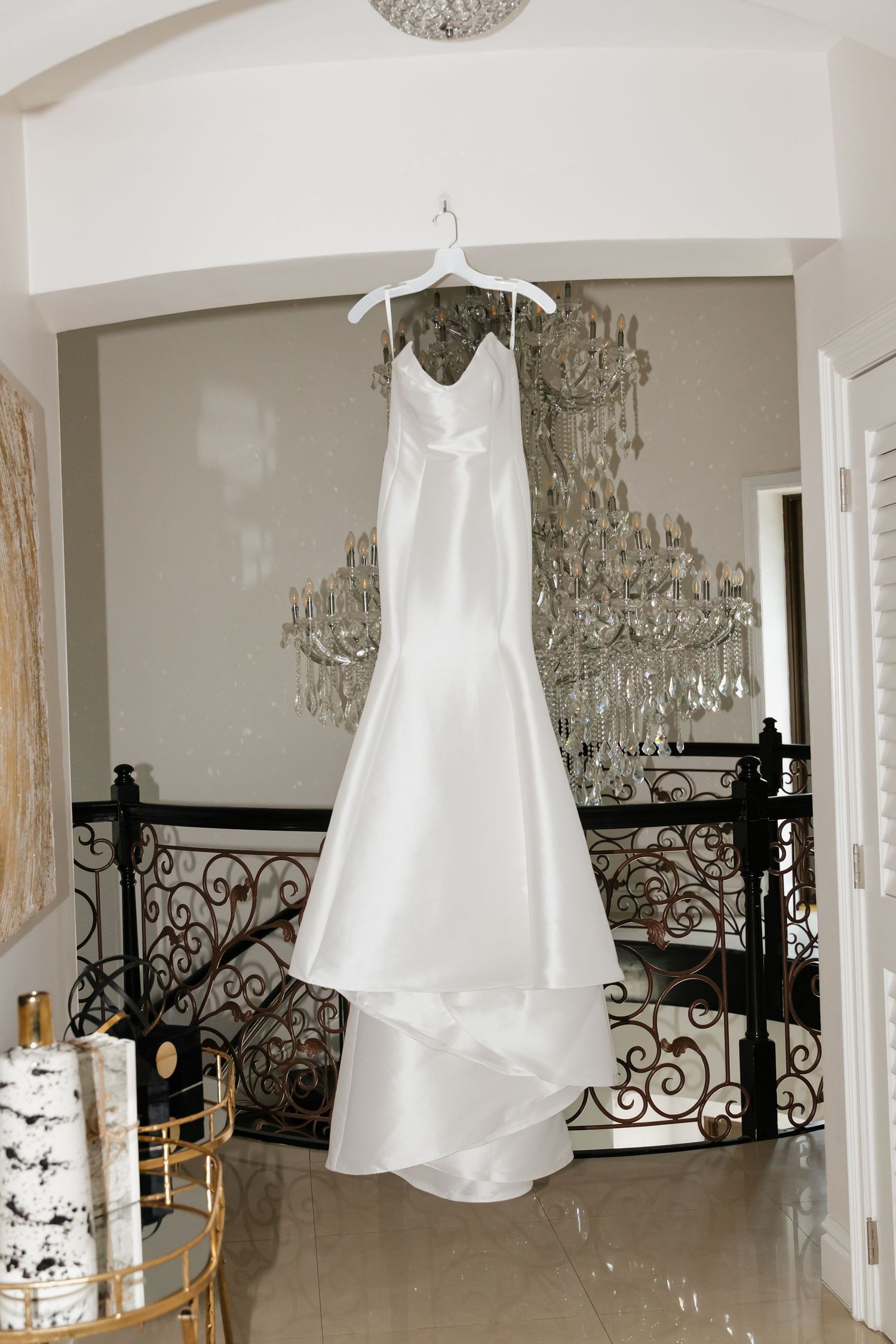 Elegant white satin mermaid wedding gown hanging on a hanger in a luxurious venue with crystal chandeliers.