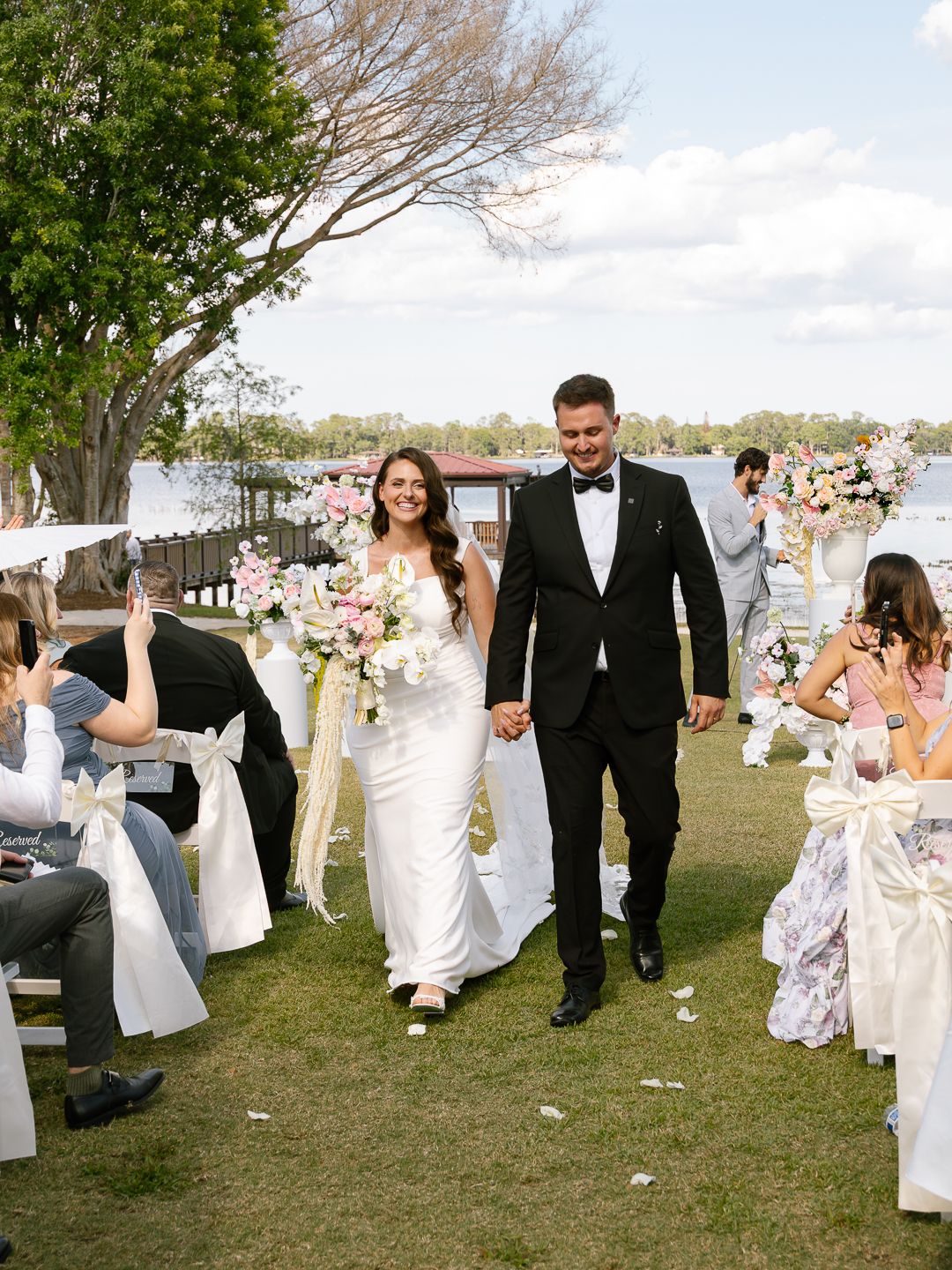 Newlyweds walk joyfully down aisle after outdoor waterfront wedding ceremony with guests cheering.