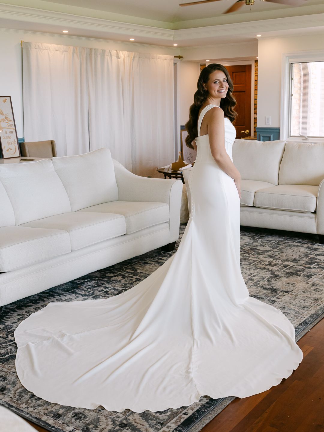 Bride in elegant white mermaid wedding gown with long train poses in bright living room, showcasing the dress's sleek silhouette.
