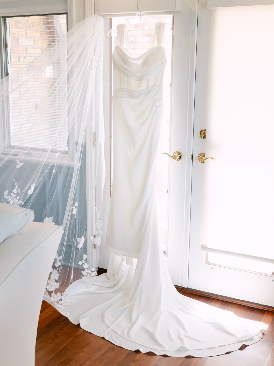 Elegant white wedding dress with long train and floral veil hanging in a sunlit doorway.