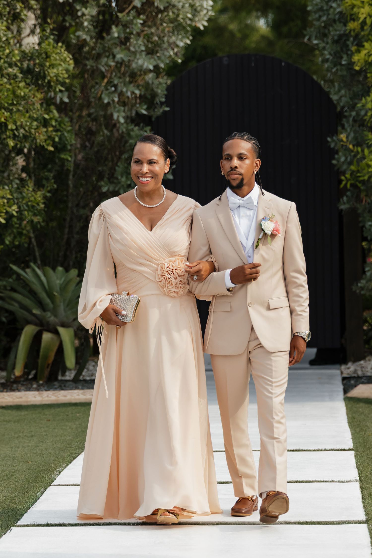 Groom in beige suit escorts woman in champagne gown down white aisle at outdoor wedding ceremony.