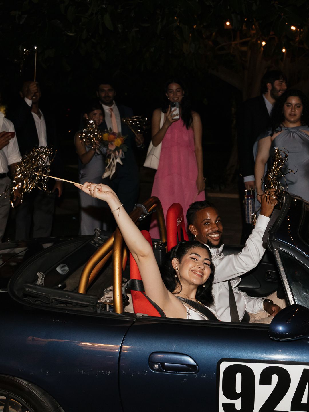 Newlyweds celebrating in vintage convertible sports car surrounded by cheering wedding guests at night.