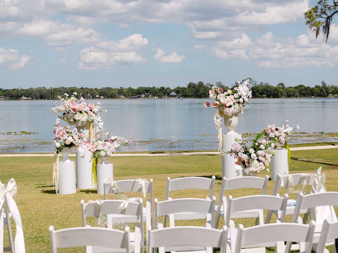 Outdoor lakeside wedding ceremony setup with white chairs and tall floral arrangements of pink and white blooms.