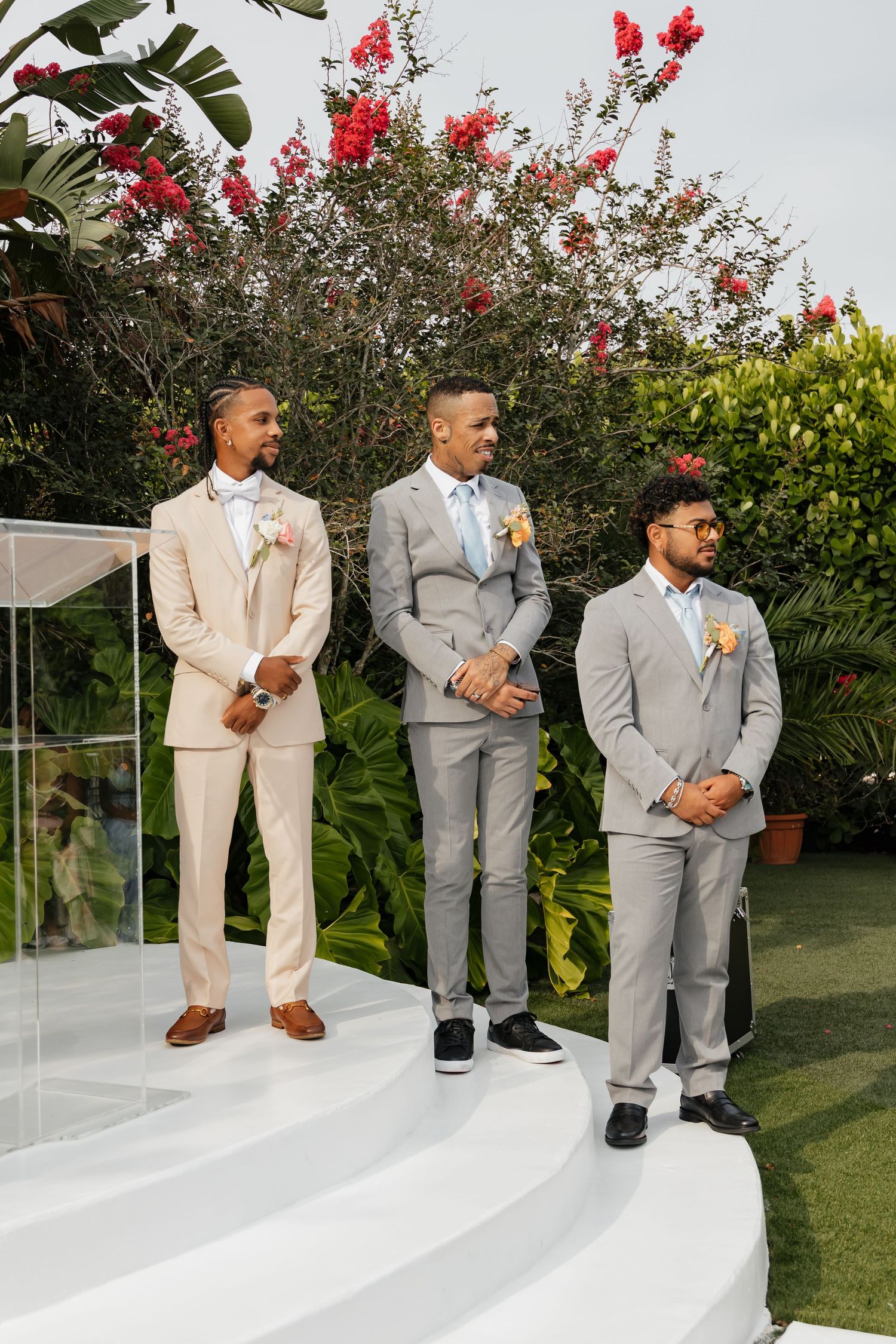 Three groomsmen in gray and beige suits standing at outdoor wedding altar surrounded by tropical flowers.