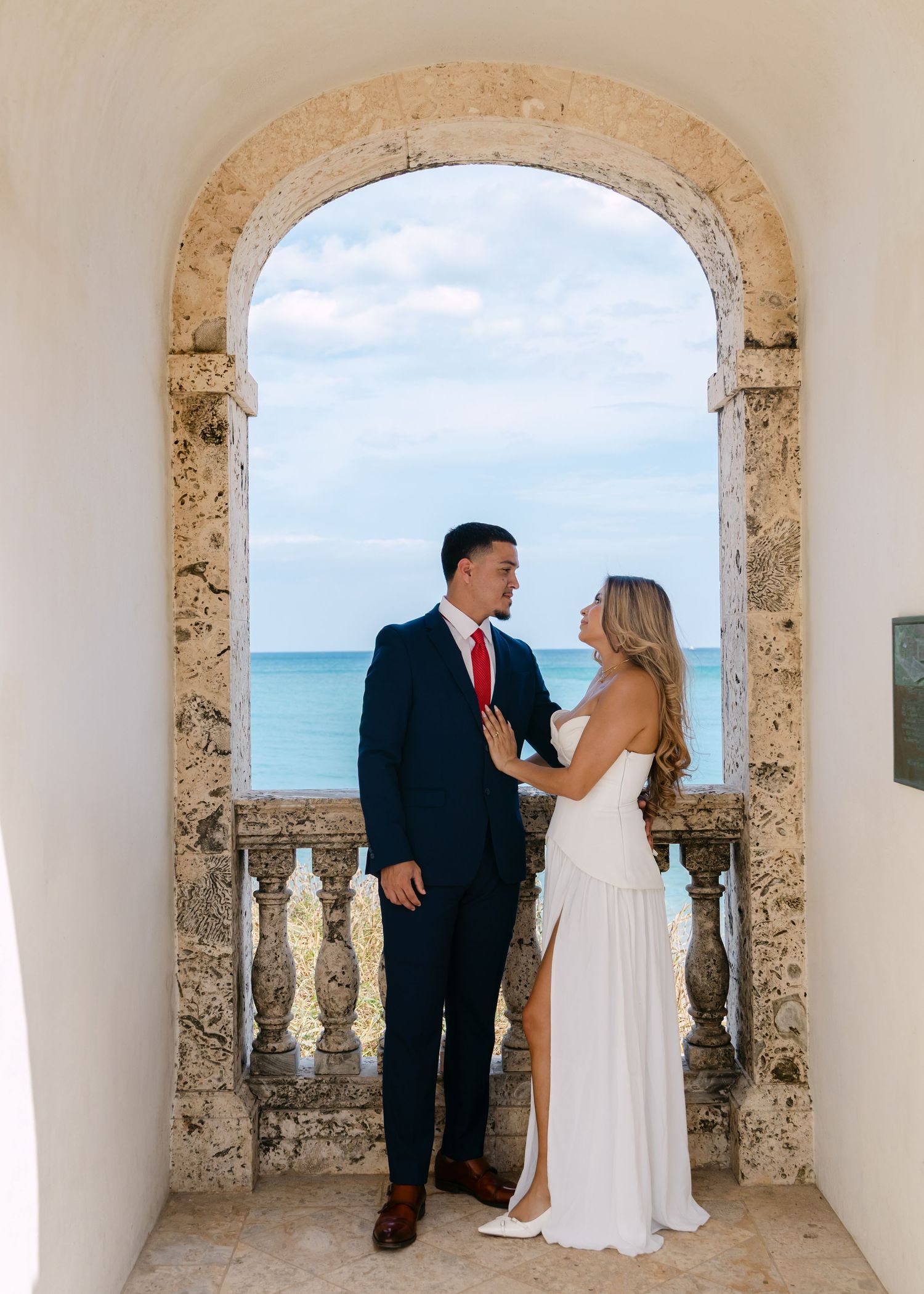 Couple in wedding attire sharing a romantic moment under a stone arch balcony overlooking the ocean.